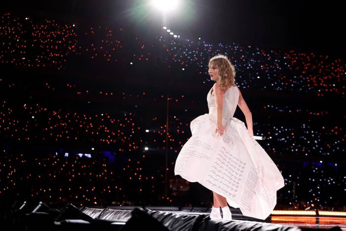 Taylor Swift in an elegant gown with text, performing onstage, surrounded by a sea of twinkling lights in a large stadium