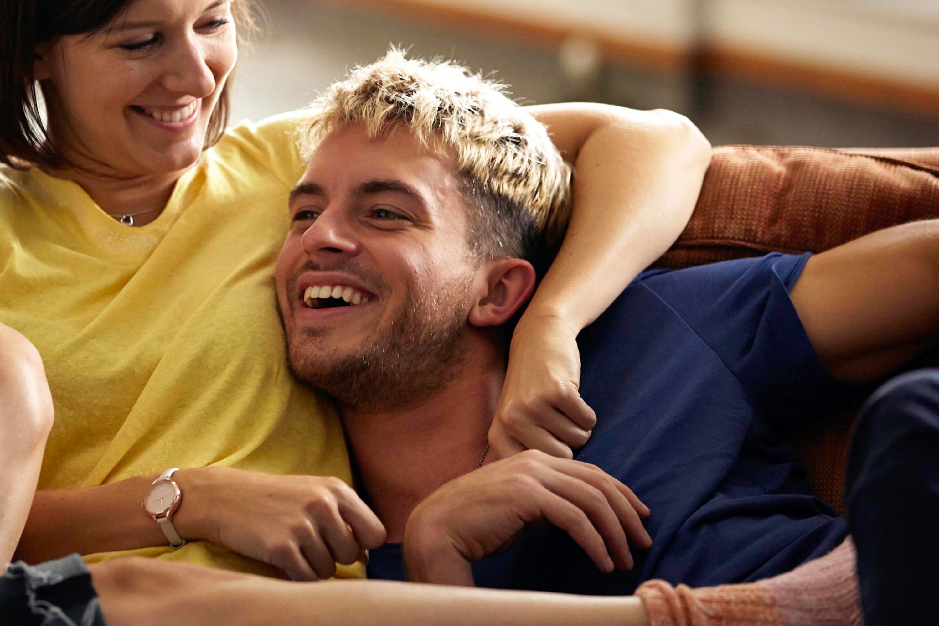Two people sitting on a couch, smiling and relaxing together. One has short hair and is wearing a yellow top, and the other has slightly longer hair, wearing a blue top