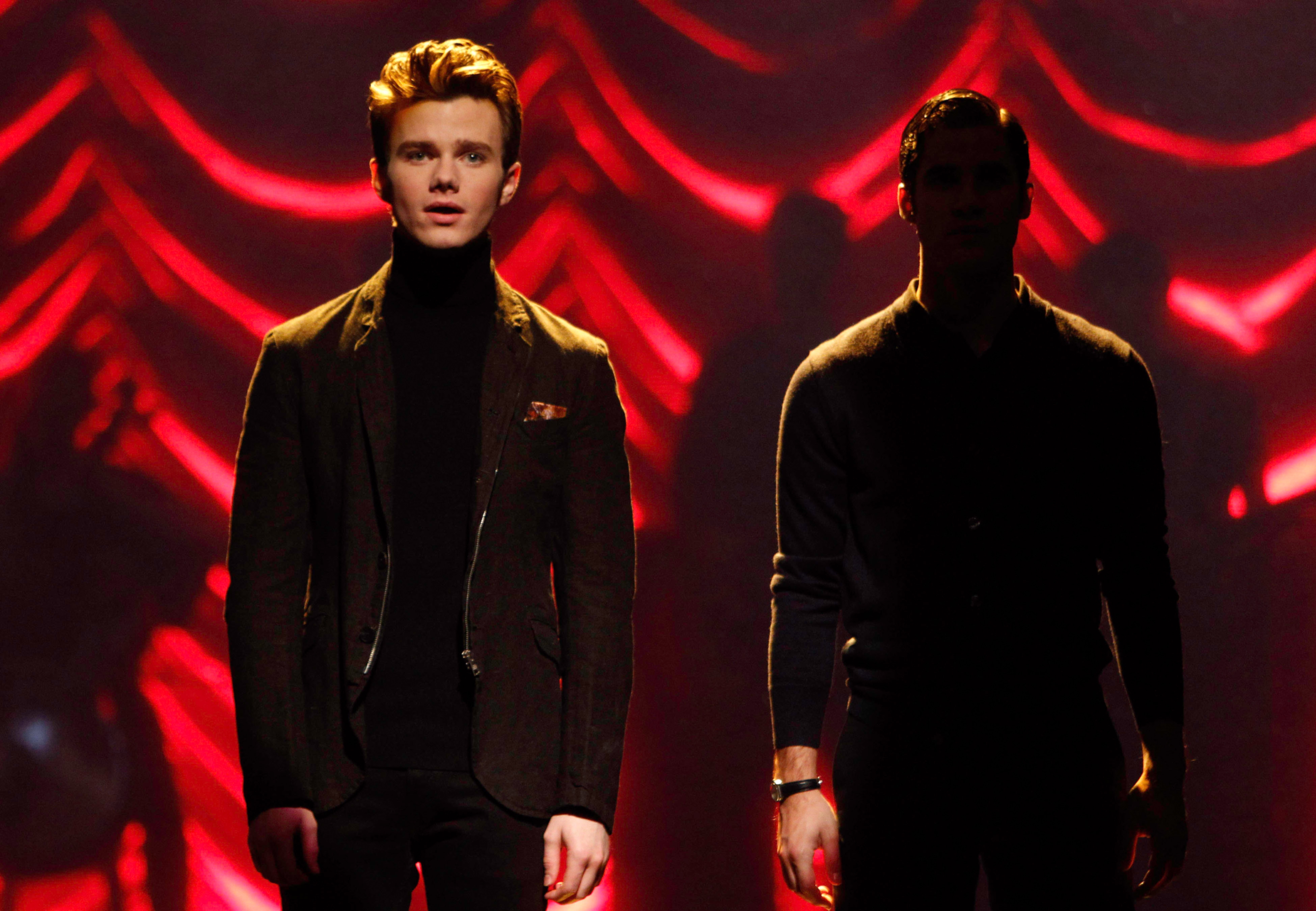 Two people stand on stage, one in a dark suit and turtleneck lit dramatically; the other is in shadow, blending with the background