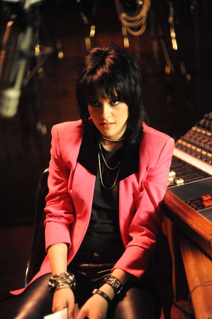 Person with short black hair in a music studio wearing a bold blazer, layered necklaces, and bracelets, with a contemplative expression