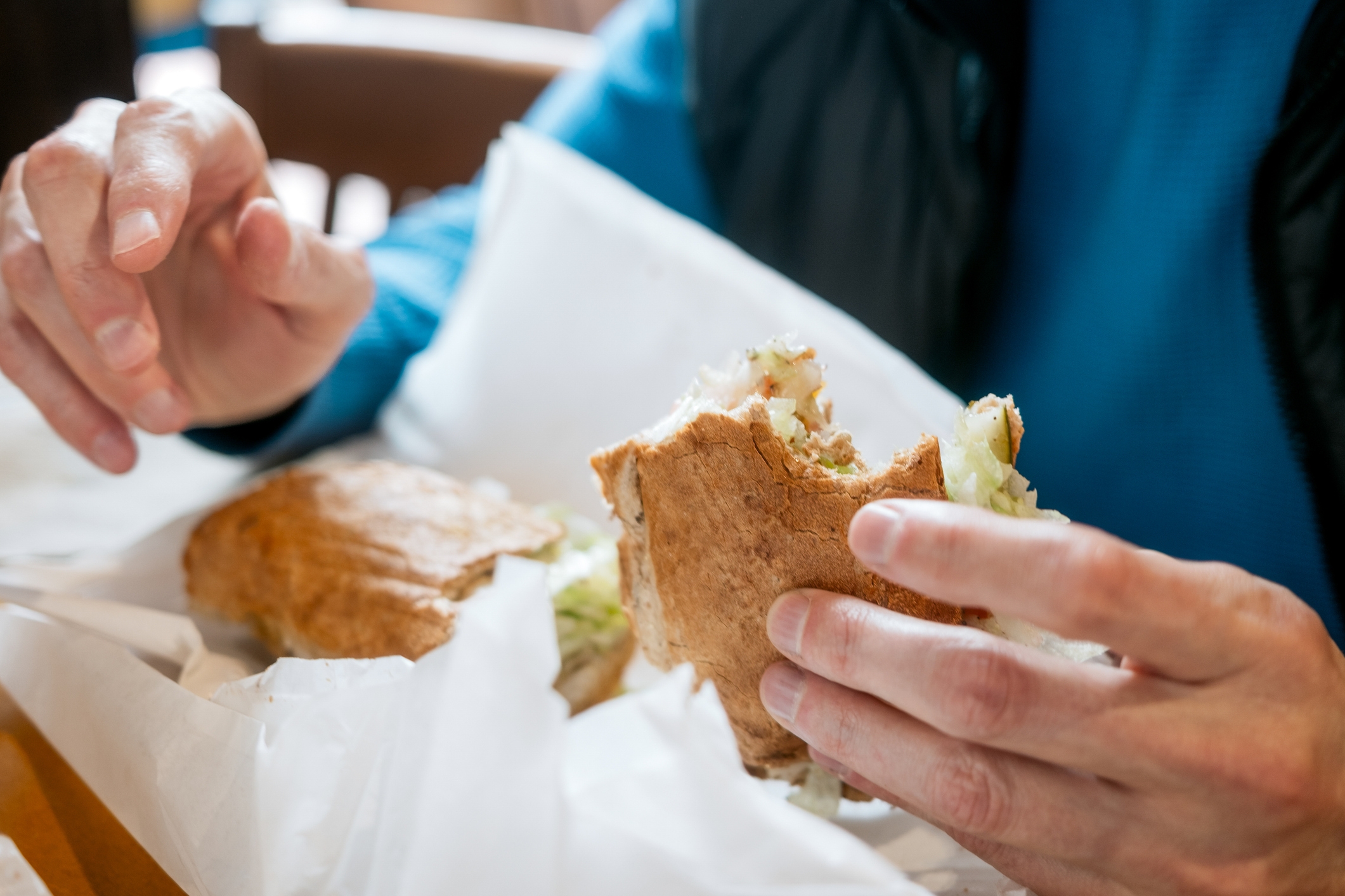 Personen som holder halvspist sandwich pakket inn i papir, med synlige ingredienser som salat og et brunt brødutsikt. Hender plassert midt-action