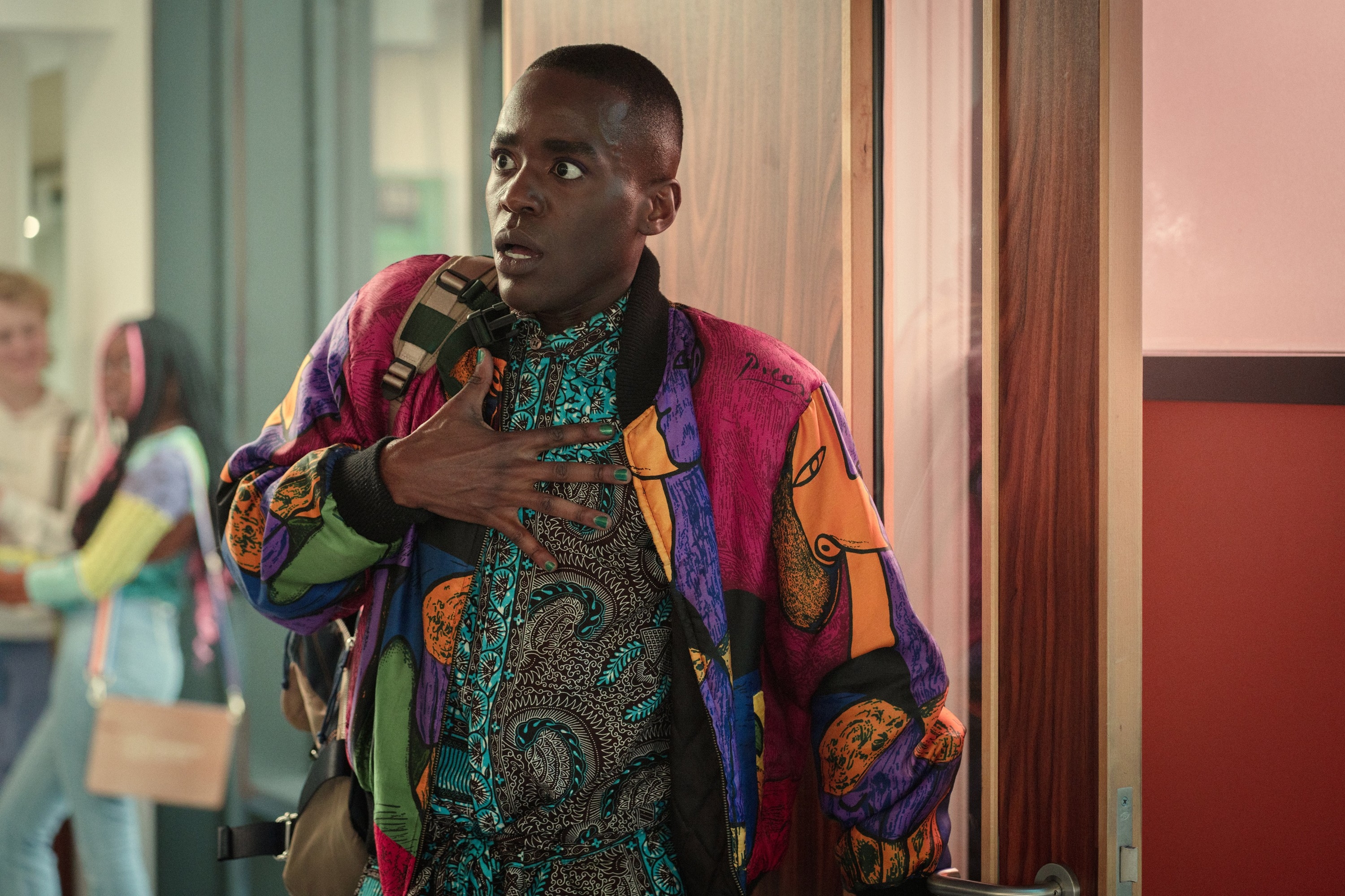 Person in a vibrant, patterned jacket with a surprised expression holds a hand to their chest, standing in a hallway setting