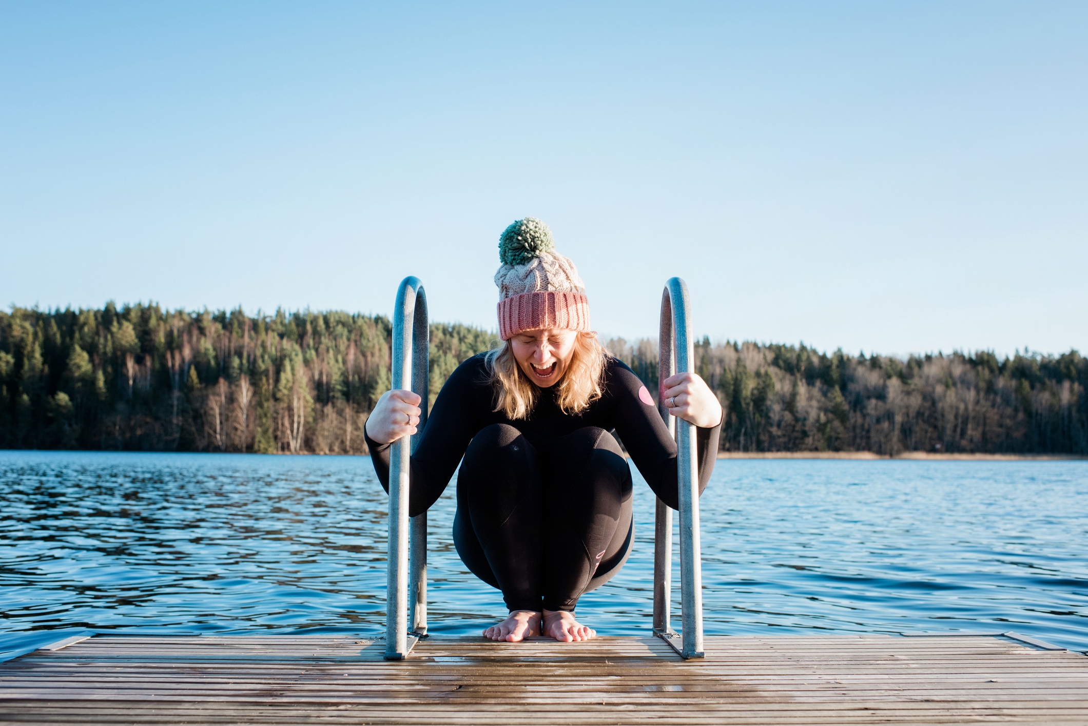 Person i en strikket hatt og våtdrakt huker seg på en innsjø dock og holder på rekkverk, med en skogkledd strandlinje i bakgrunnen