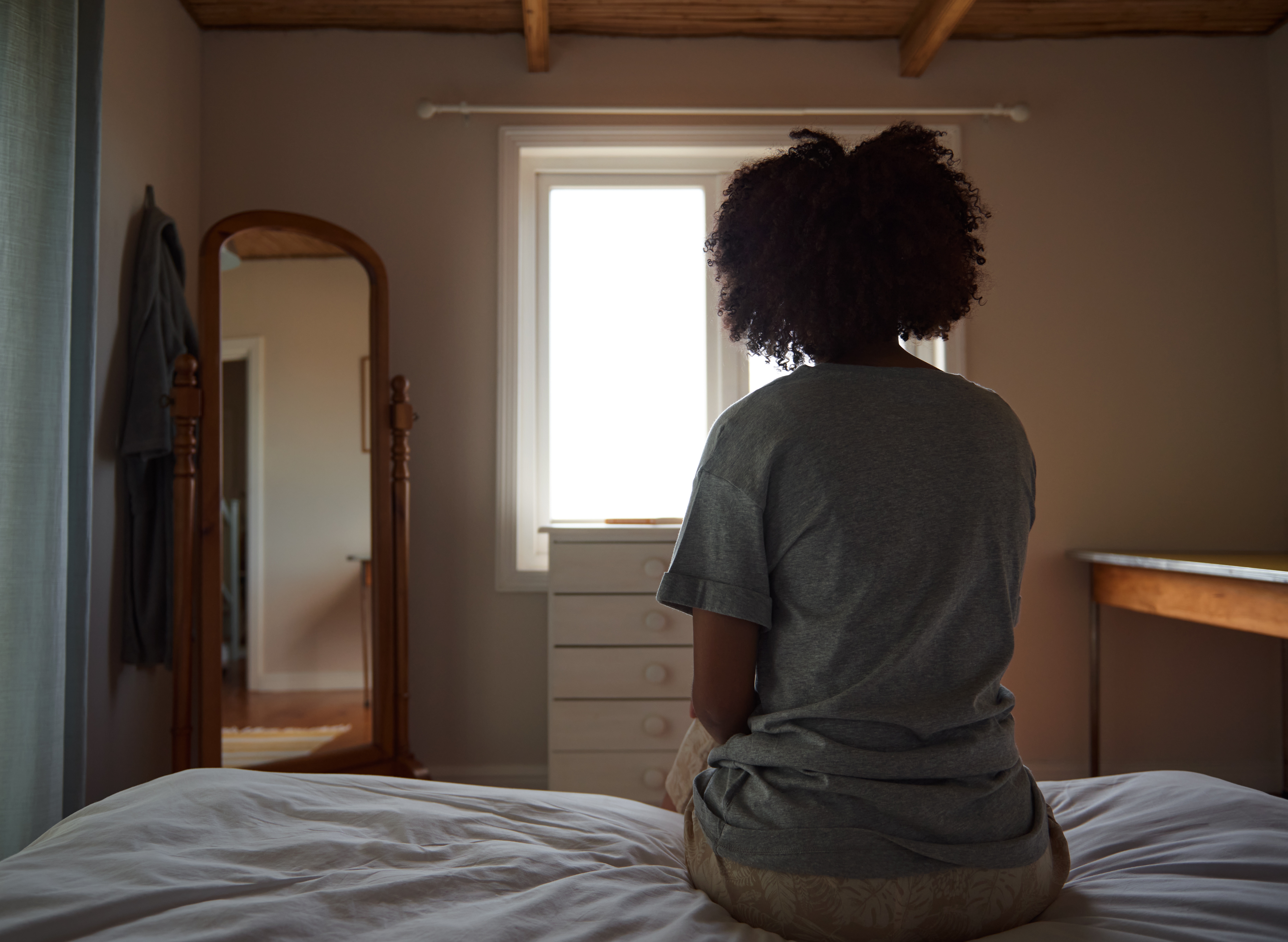 Person som sitter på en seng mot et vindu, med naturlig lys som kommer inn, ved siden av et stående speil og en kommode