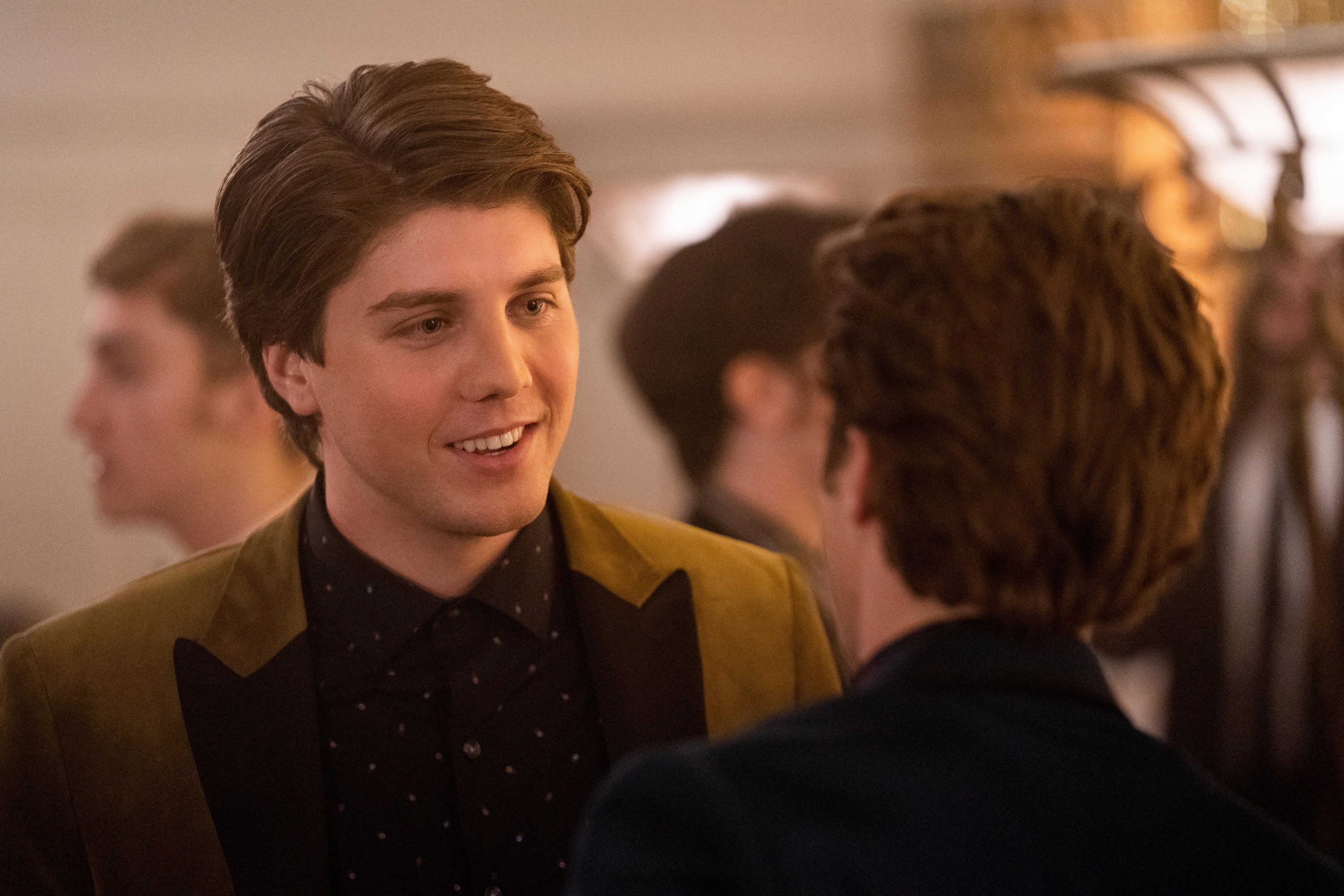 Two people are engaged in conversation at a social event. The person facing the camera is wearing a dark shirt and a blazer