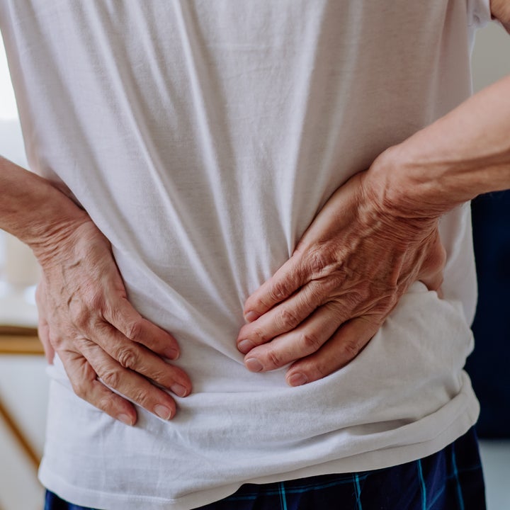 Person holding their lower back, indicating discomfort or pain, standing in a bedroom setting