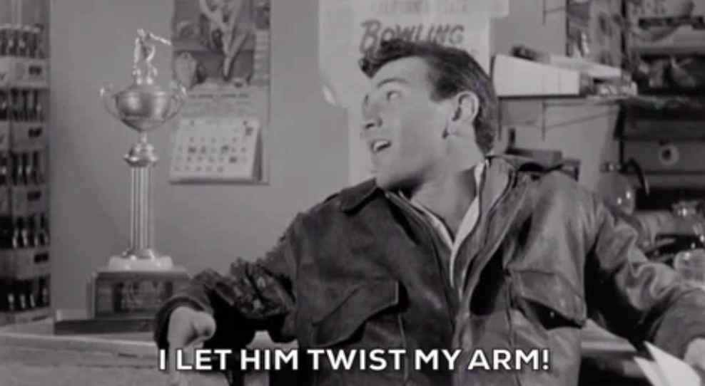 A person in a leather jacket smiles, saying, "I let him twist my arm!" in a room with a trophy on a shelf behind them