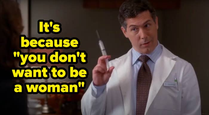 A doctor in a lab coat holds a syringe, speaking to a person off-camera in an office setting. A lamp and flowers are visible in the background