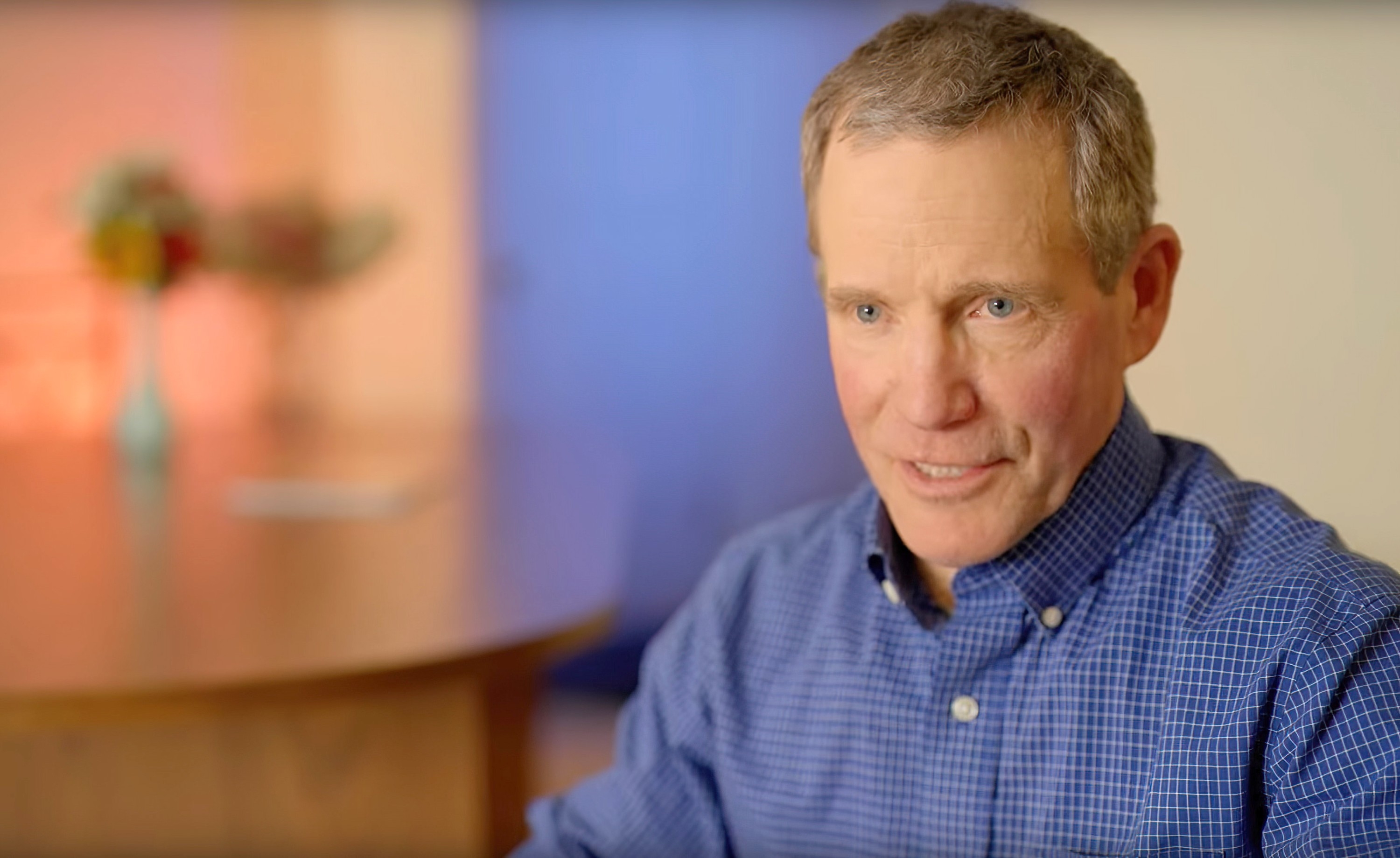 Man in a checkered shirt is sitting indoors, looking to the side of the camera. Blurred background with table and vase