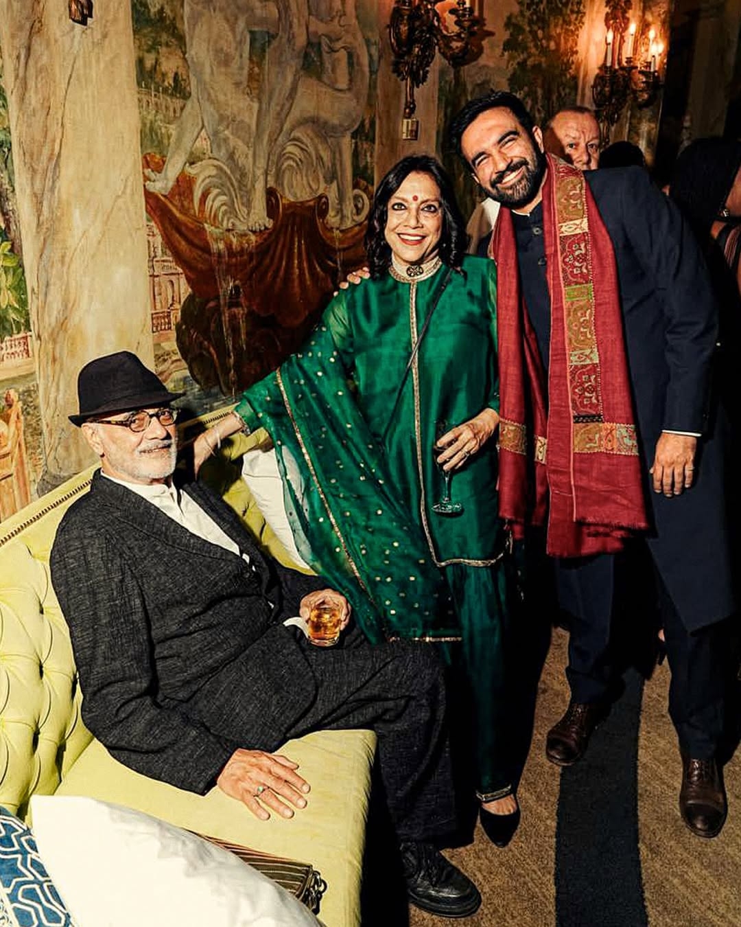 Three people are joyfully posing at a social event. A seated man in a suit and hat holds a drink. Two standing guests wear elegant, traditional attire