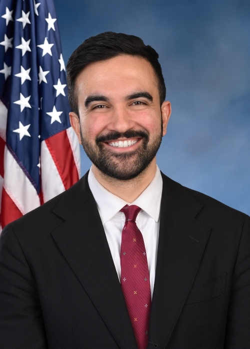 A smiling person in a suit stands in front of a U.S. flag, set against a neutral backdrop