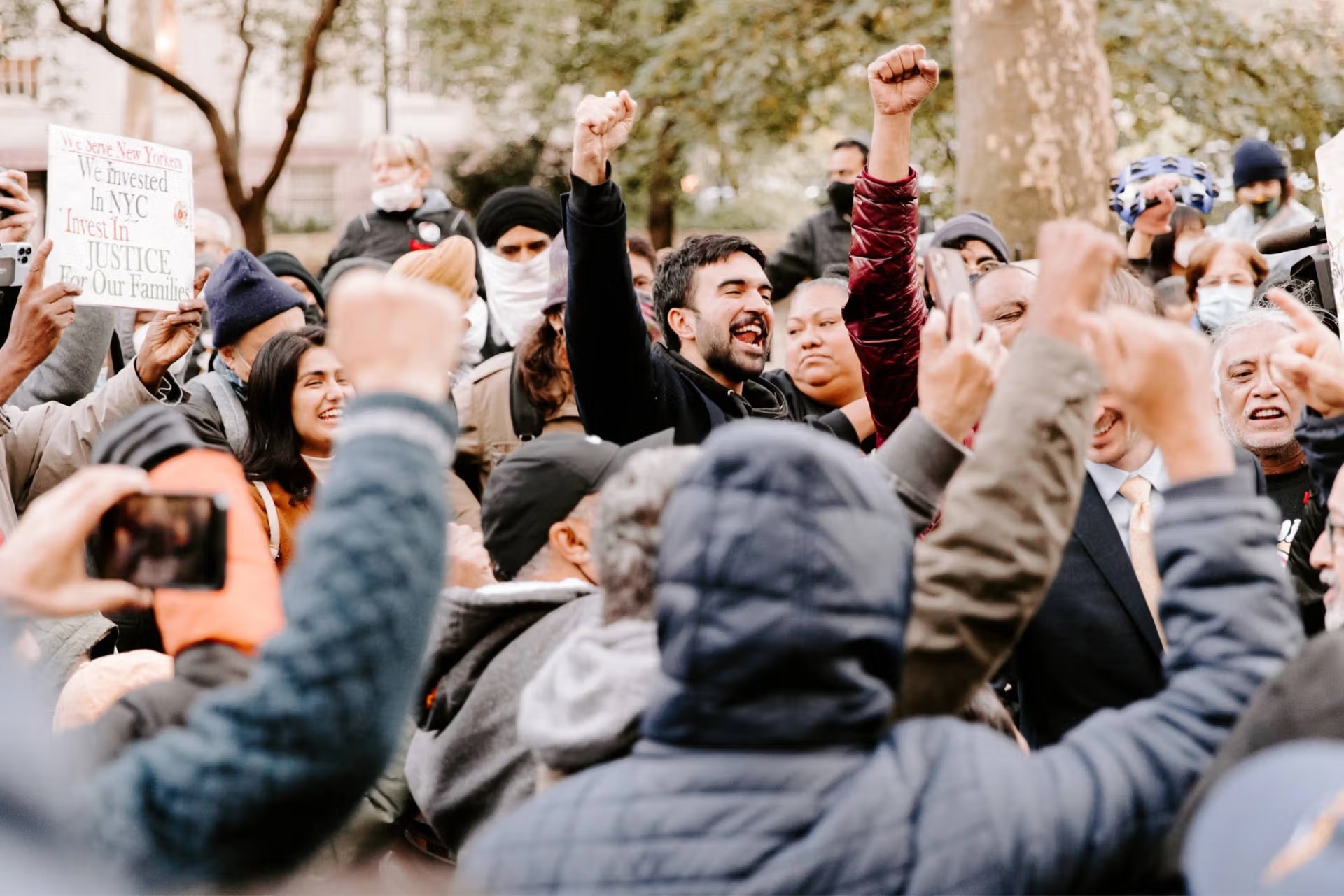 A diverse crowd gathers outdoors in a spirited protest, many raising fists in solidarity. A man at the center raises his fist enthusiastically