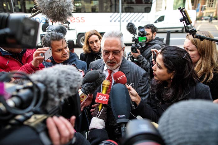 A man in a suit is surrounded by reporters with microphones and cameras, giving a statement amidst a flurry of media activity