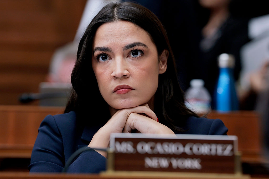 A person with long hair, looking serious, rests their chin on crossed hands at a formal meeting. A nameplate reading "Ms. Ocasio-Cortez, New York" is visible