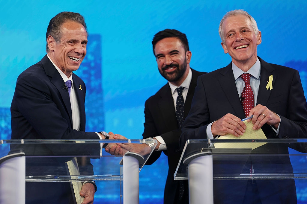 Three people in suits stand at podiums, smiling and shaking hands, each wearing a yellow ribbon