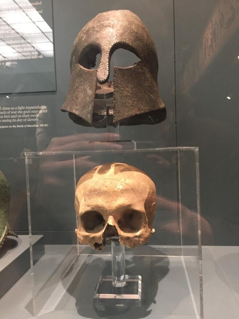 Ancient metal helmet displayed above a human skull in a museum exhibit