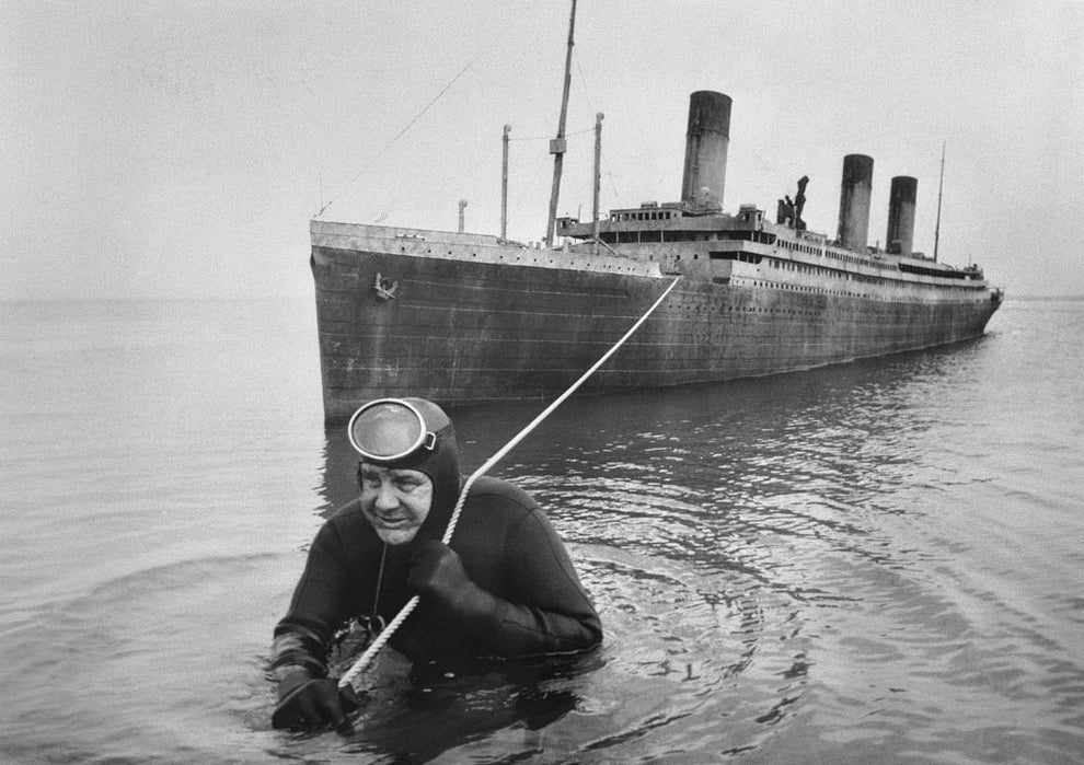 A diver wades in water holding a stick, with a large vintage ship in the background, resembling an iconic ocean liner
