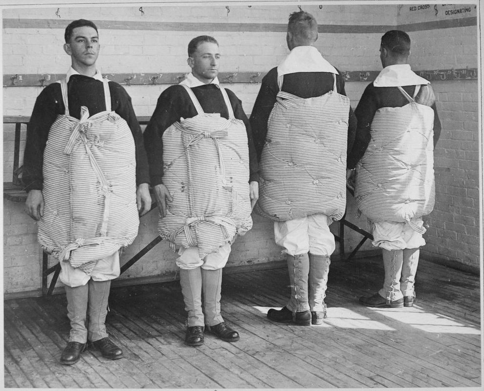 Four men in a locker room wearing striped padded suits for experimental football gear testing, likely from the early 20th century
