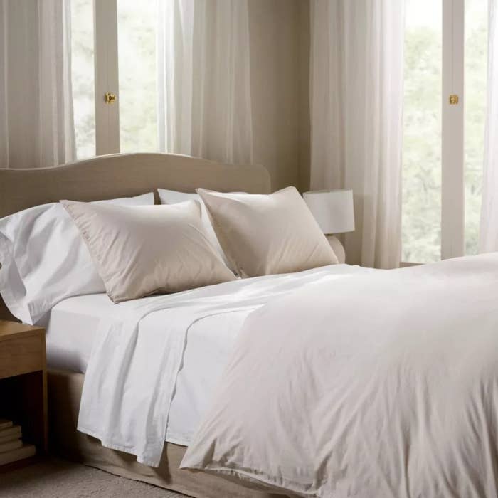 A neatly made furniture with fluffy pillows and a woody headboard is adjacent ample windows with sheer curtains, creating a serene, well-lit chamber atmosphere