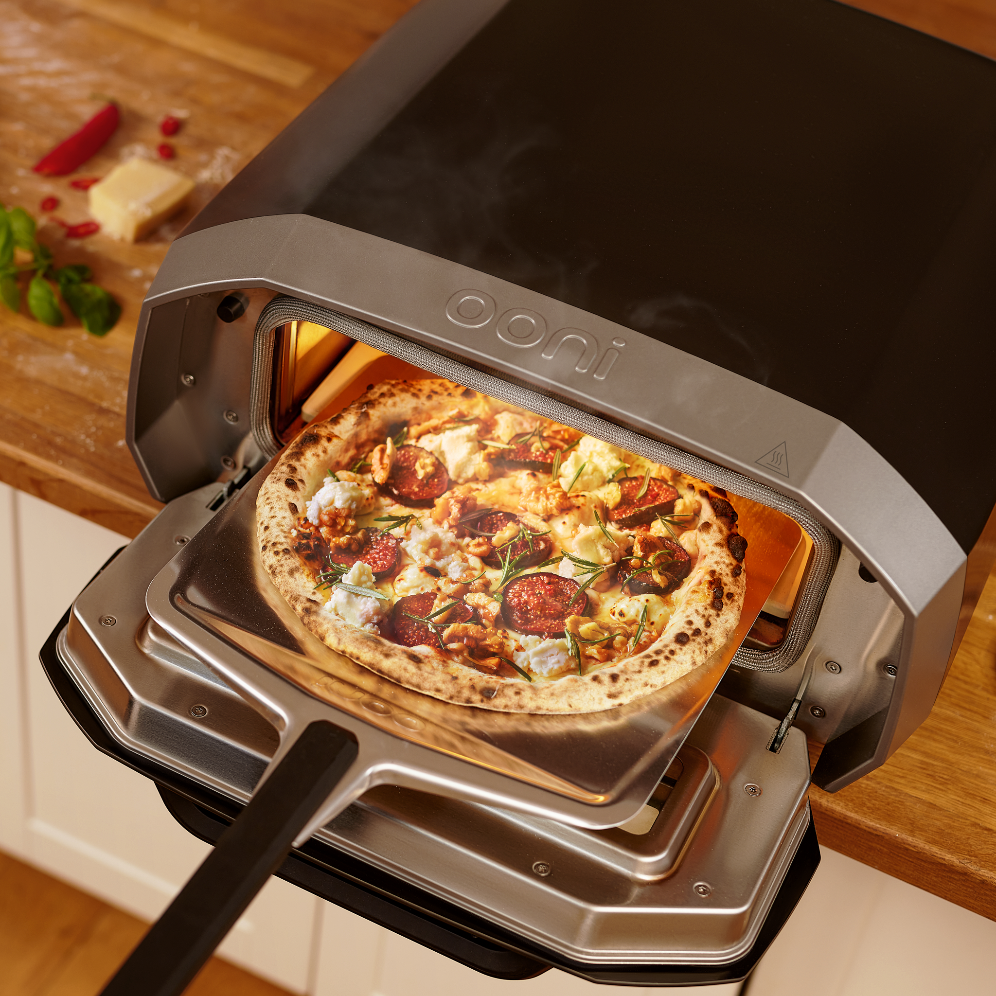 Pizza cooking in a portable, home pizza oven on a kitchen counter, with ingredients like cheese and herbs nearby