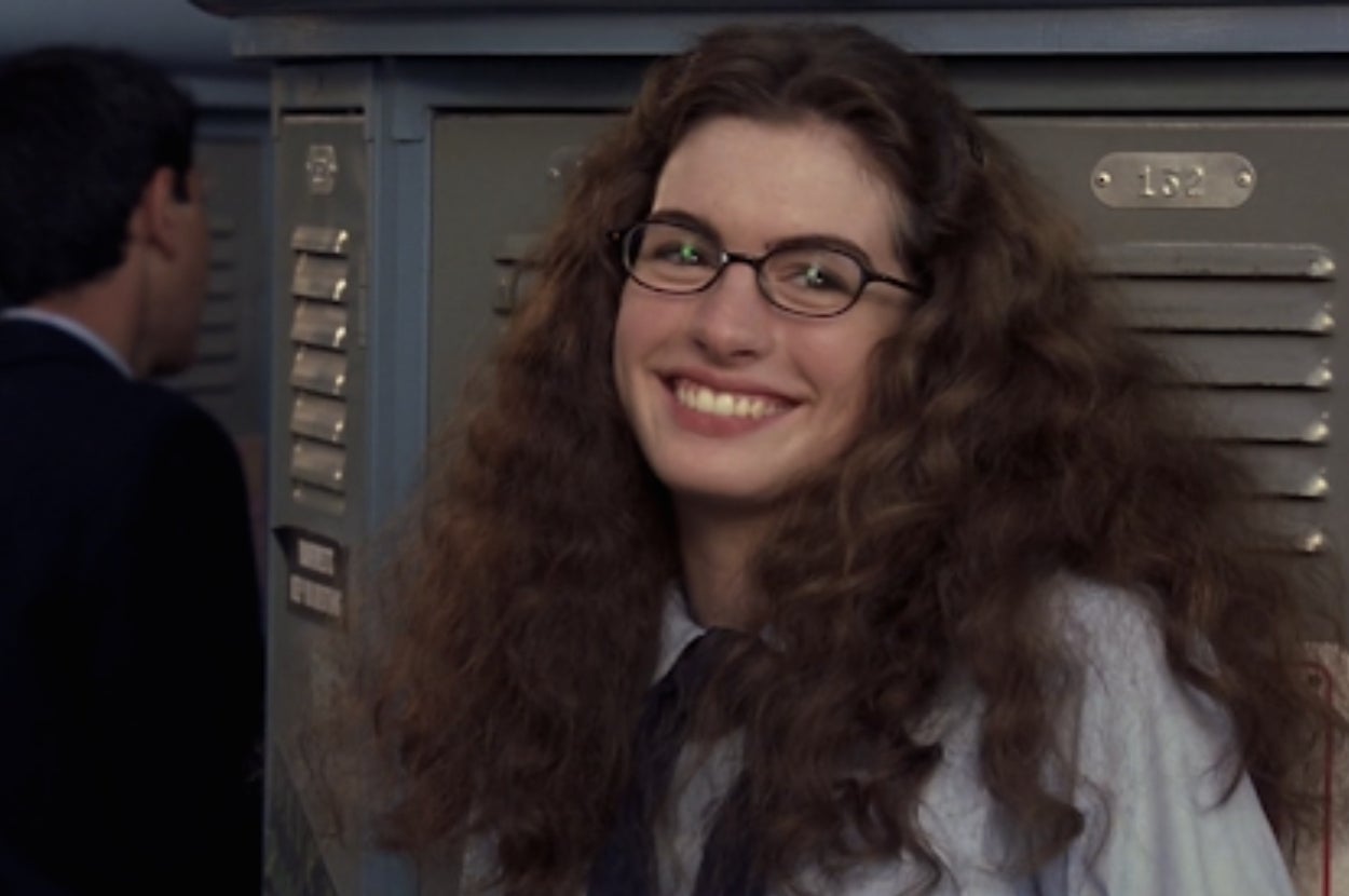 Person with curly hair and glasses smiles in front of lockers, wearing a button-up shirt and tie