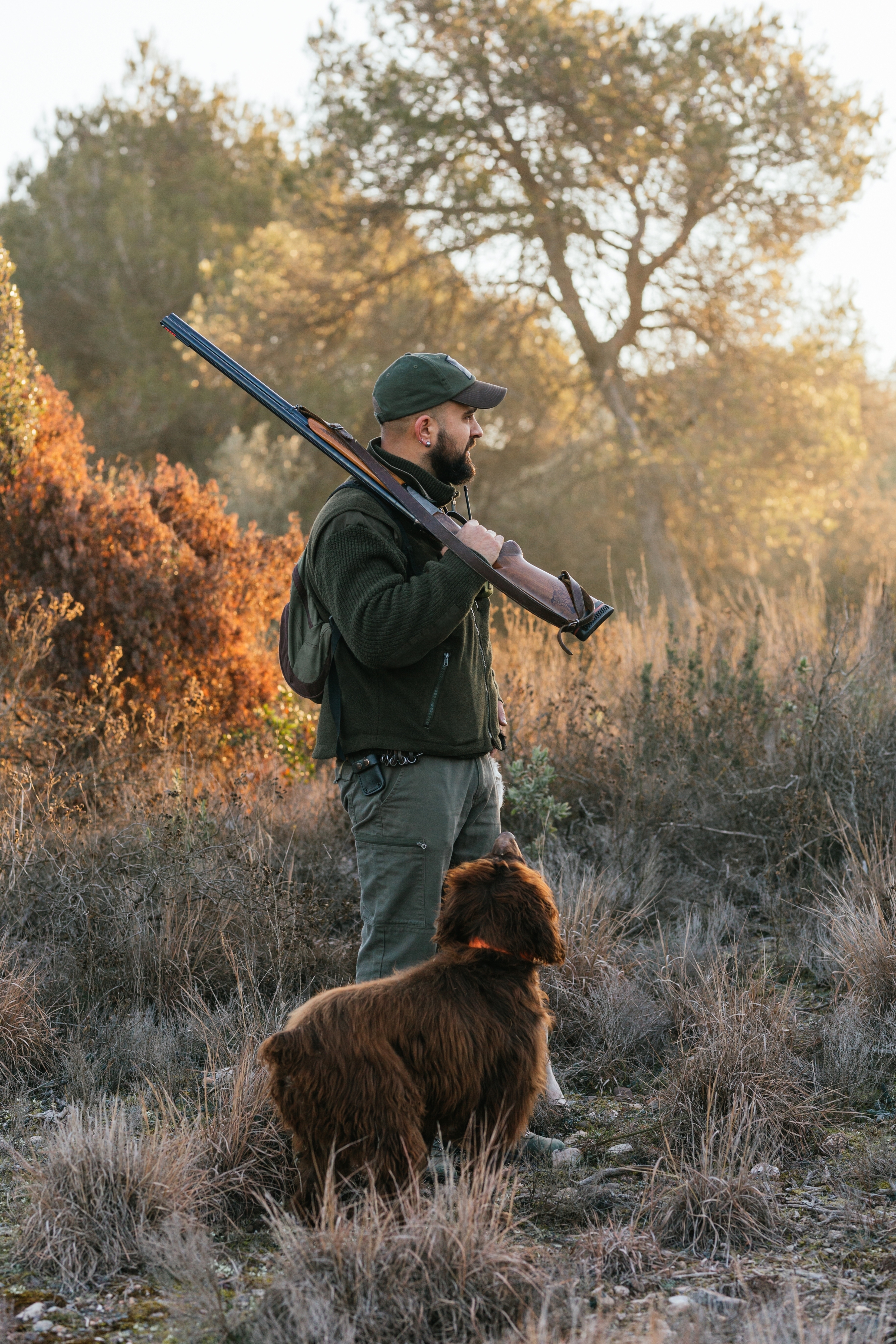 A person in outdoor gear holds a rifle, standing in a field with a fluffy dog, surrounded by trees and tall grass