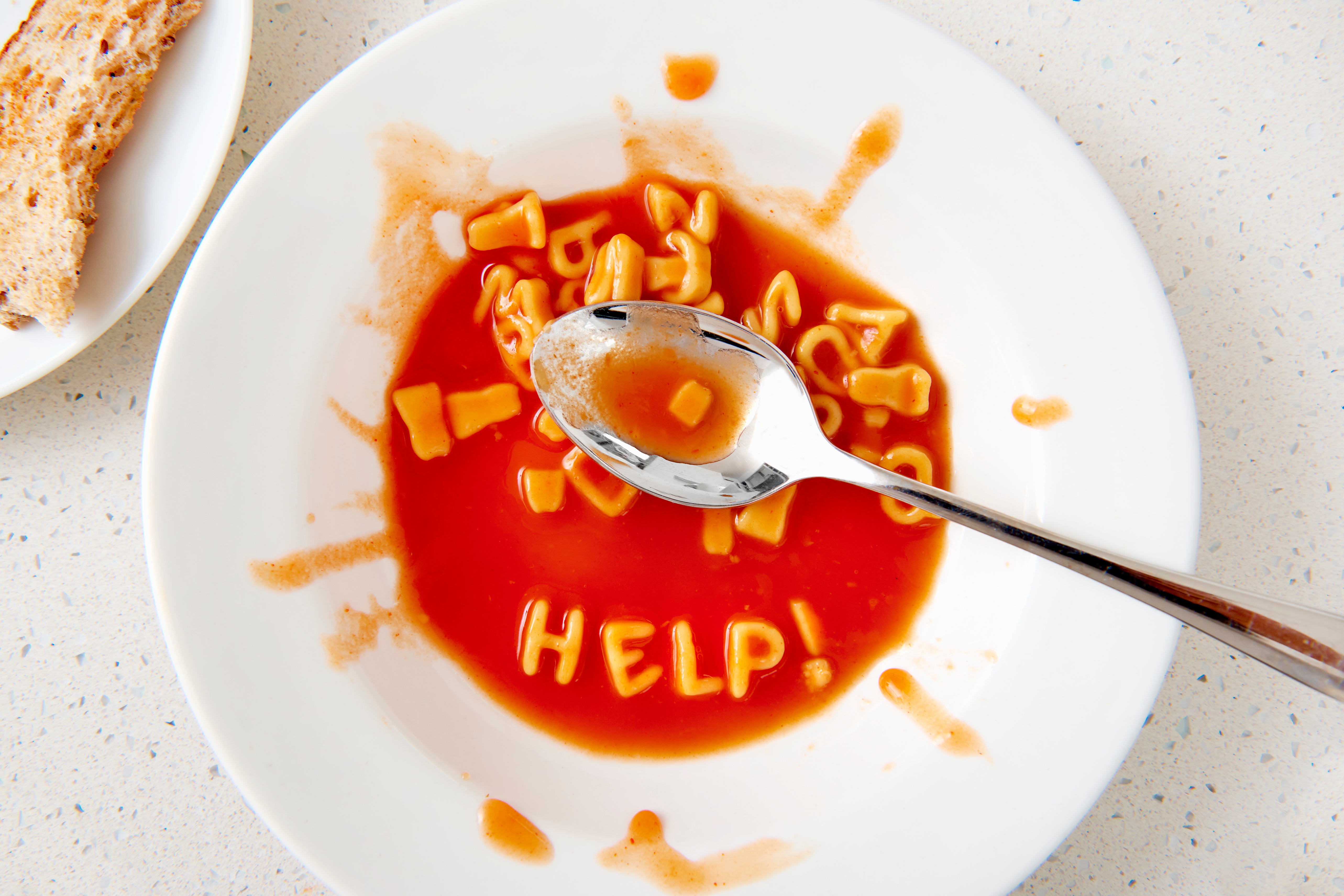 Alphabet pasta in tomato soup on a white plate spells "HELP!" with a spoon nearby. Toast is visible on the side