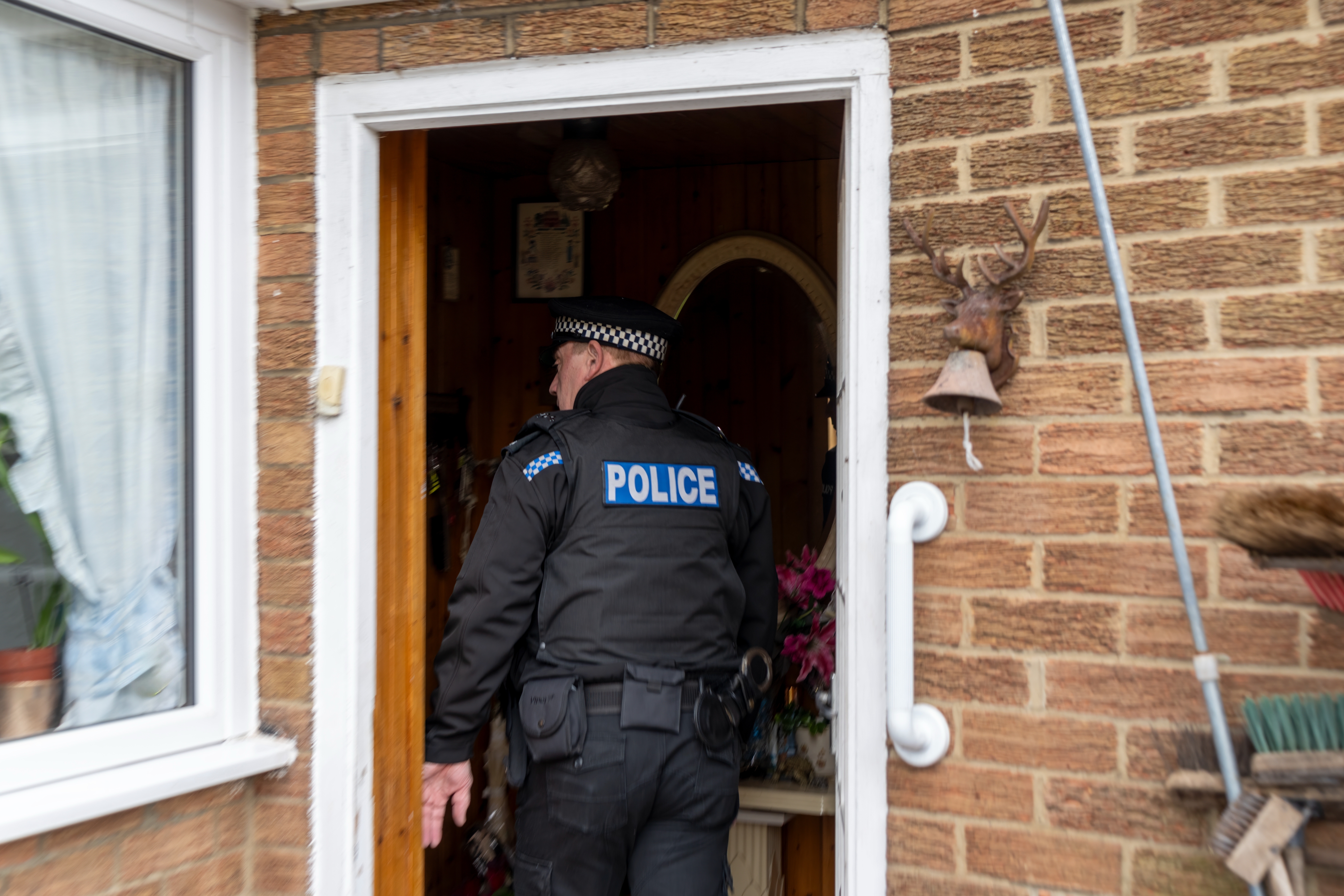 Police officer enters a brick house through an open door, wearing a uniform and hat