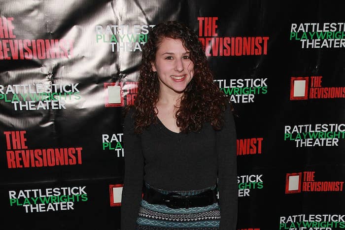 Person smiling at theater event with curly hair, wearing a knit top and a patterned skirt, standing in front of a backdrop with play text