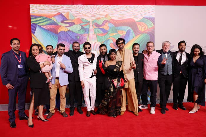 Group of people on a red carpet, with some wearing formal and stylish attire. Colorful artwork serves as the backdrop