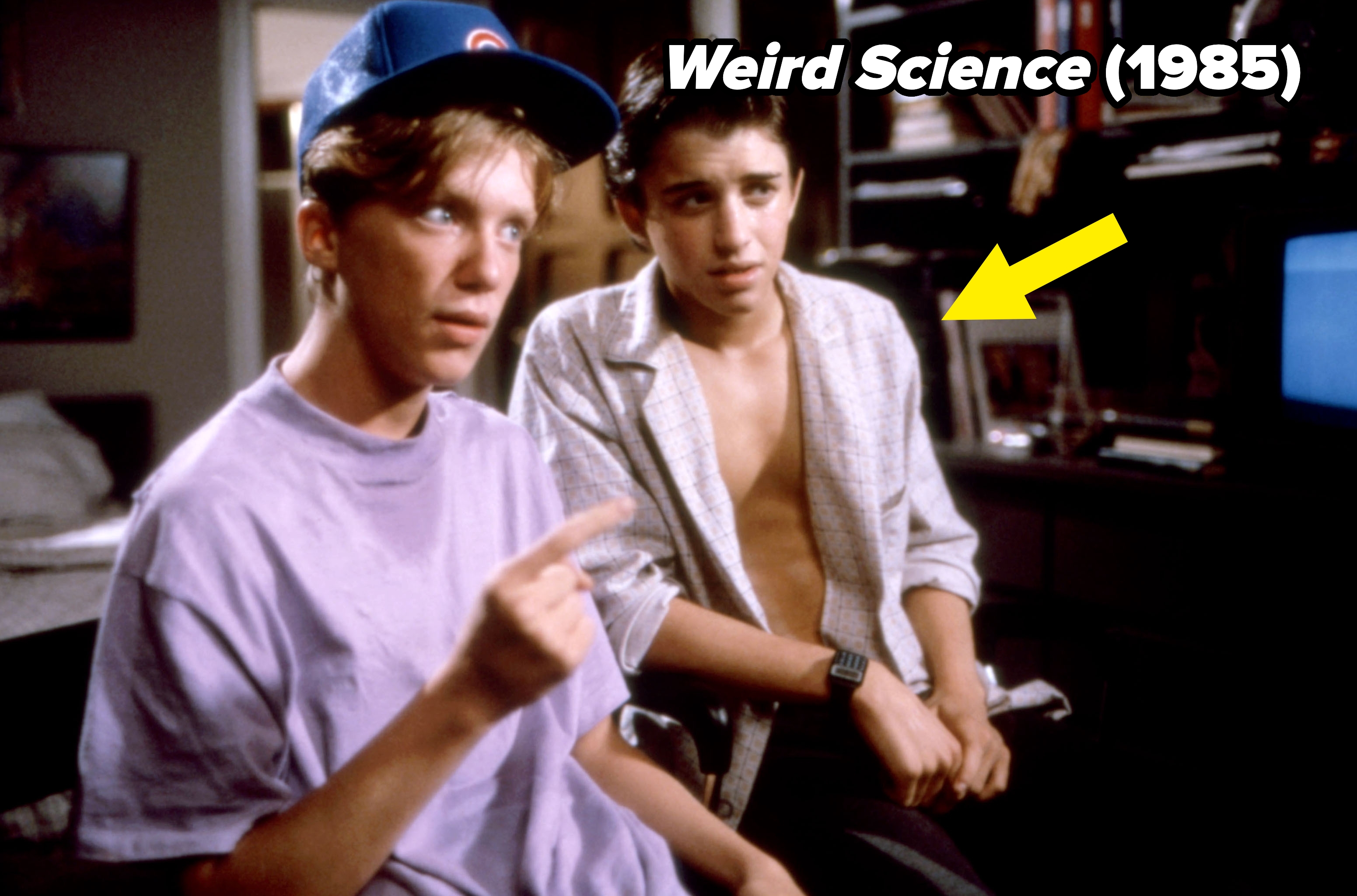 Two young men sit in a room, one wearing a T-shirt and cap, the other in an open shirt. Both look thoughtful or engaged in conversation