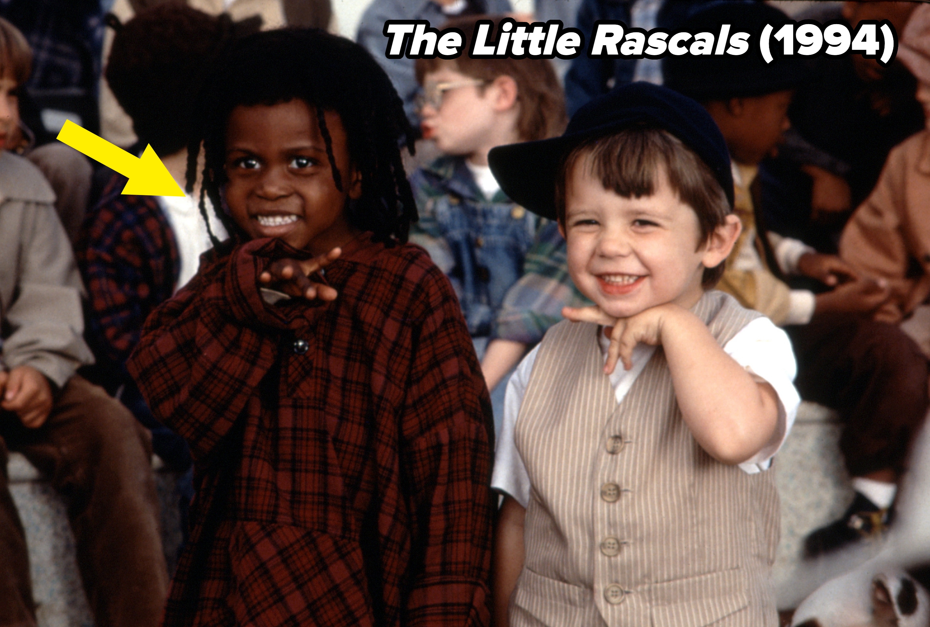 Two children smiling and playfully gesturing in a group setting. One wears a plaid shirt, the other a vest and cap