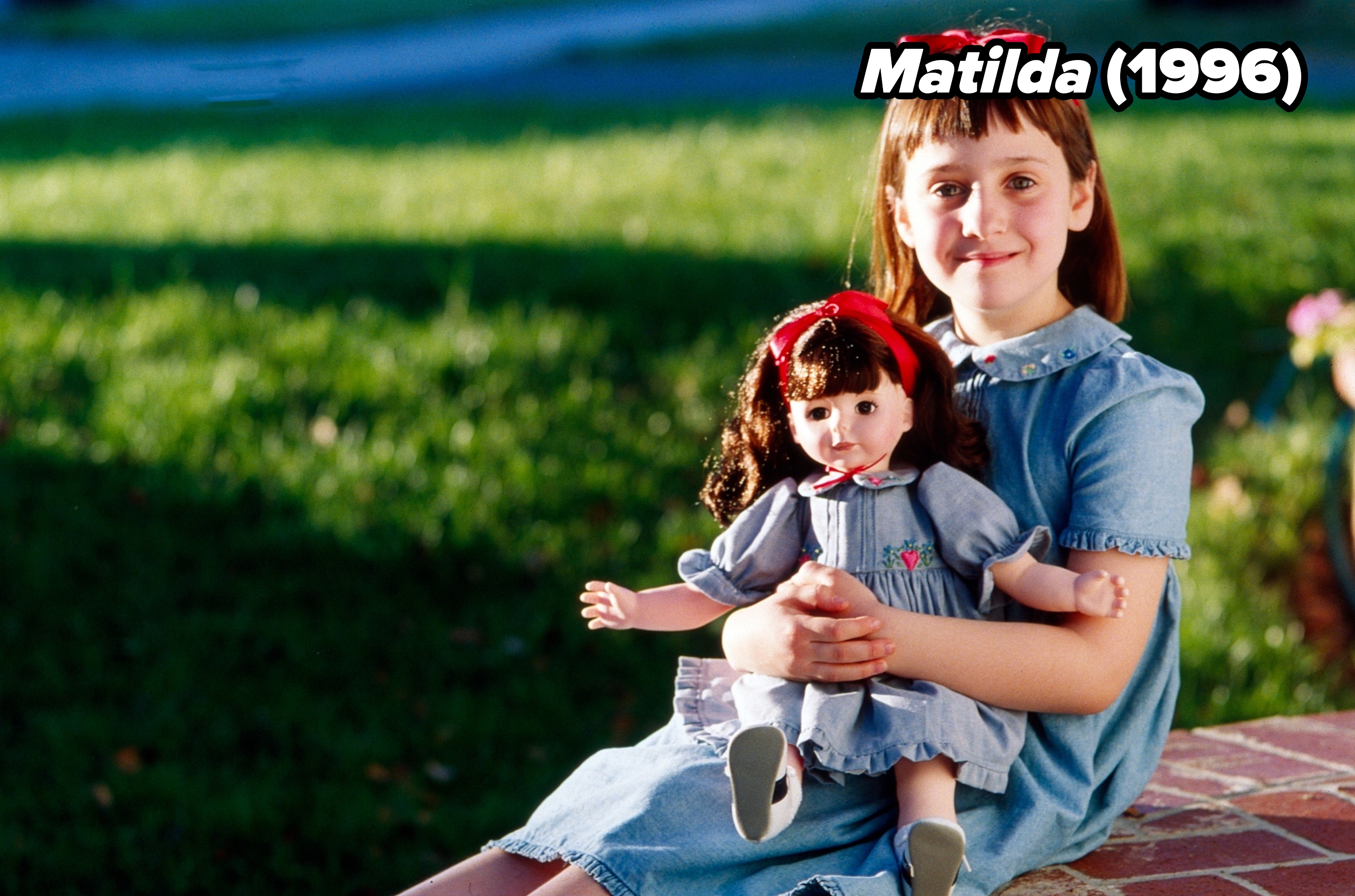 A young girl in a blue dress sits outside on a brick ledge, holding a doll dressed similarly, both with matching red bows