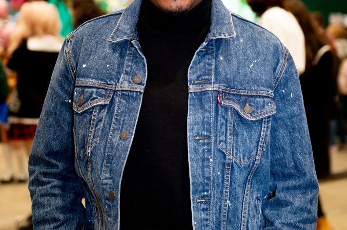 Person smiling, wearing a denim jacket over a black turtleneck at a lively event with plush toys in the background