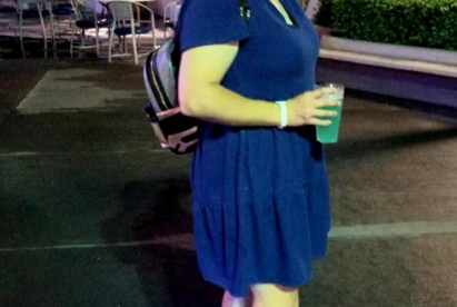 Person smiling with hand on head, wearing a casual dress and glowing shoes, holding a drink, outdoors at night