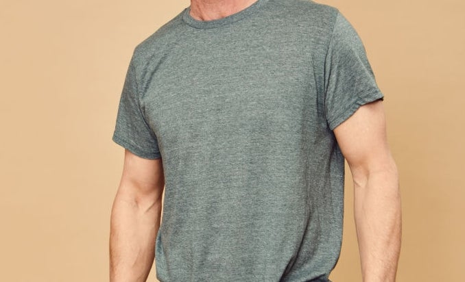 Man smiling, wearing a casual short-sleeve t-shirt and trousers, standing against a plain background