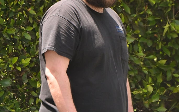 Person wearing a cap, sunglasses, a casual t-shirt, and pants stands outside by greenery