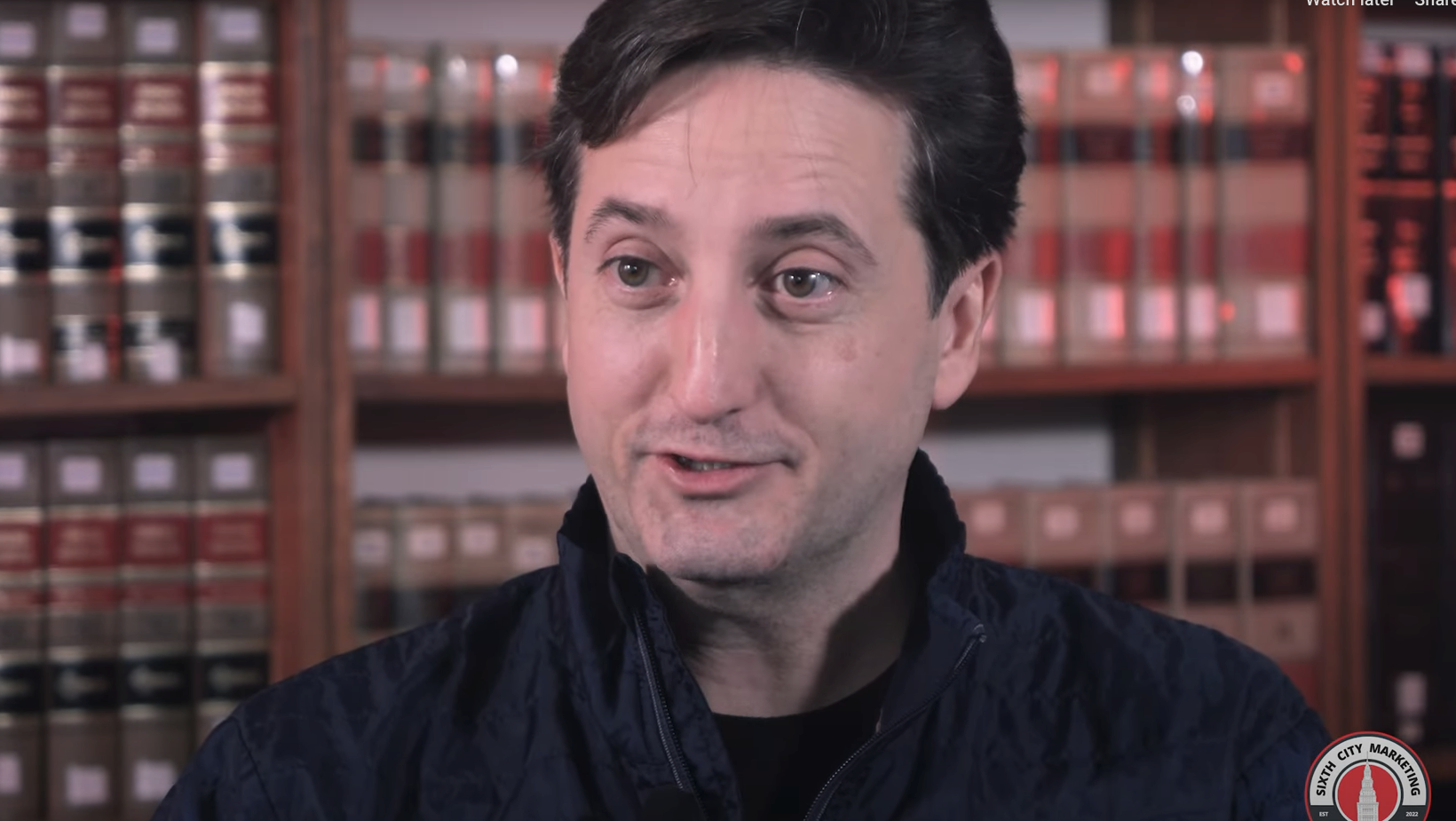 Person in jacket speaking, with bookshelves in the background