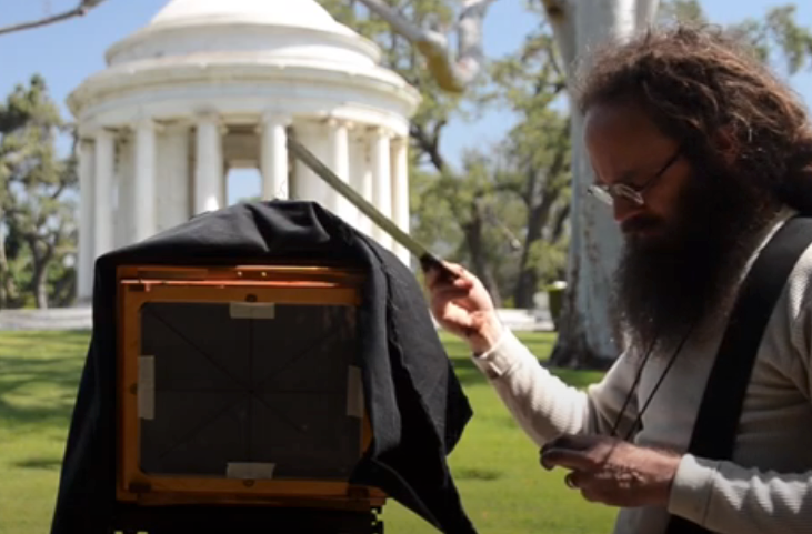 A person with long hair and a beard sets up a large-format camera in a park near a white gazebo