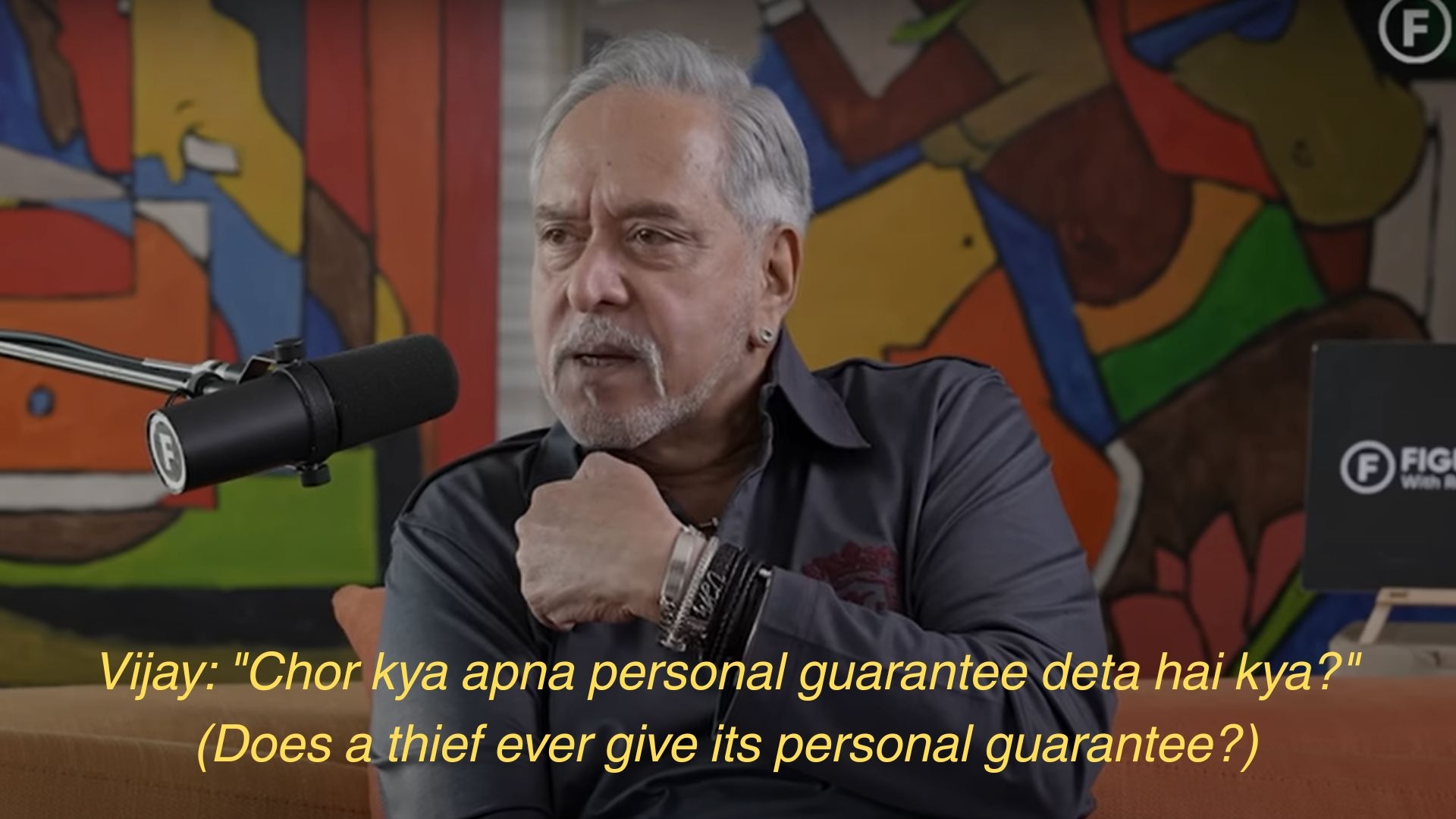 A person in a casual gray shirt speaks into a microphone, gesturing with their hand. Subtitles discuss a thief providing a personal guarantee