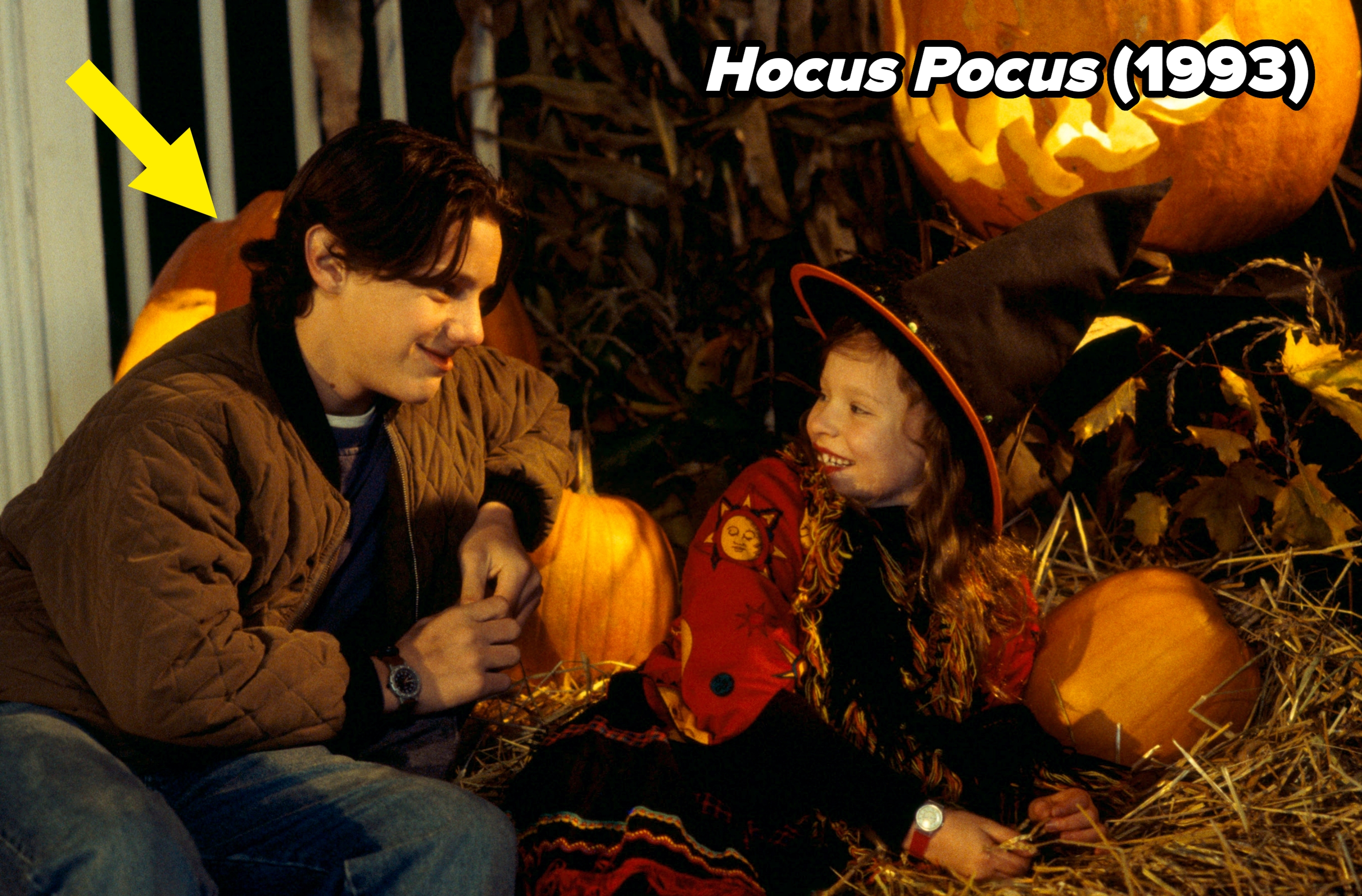A person and a child in a witch costume sit on hay surrounded by carved pumpkins, sharing a happy moment