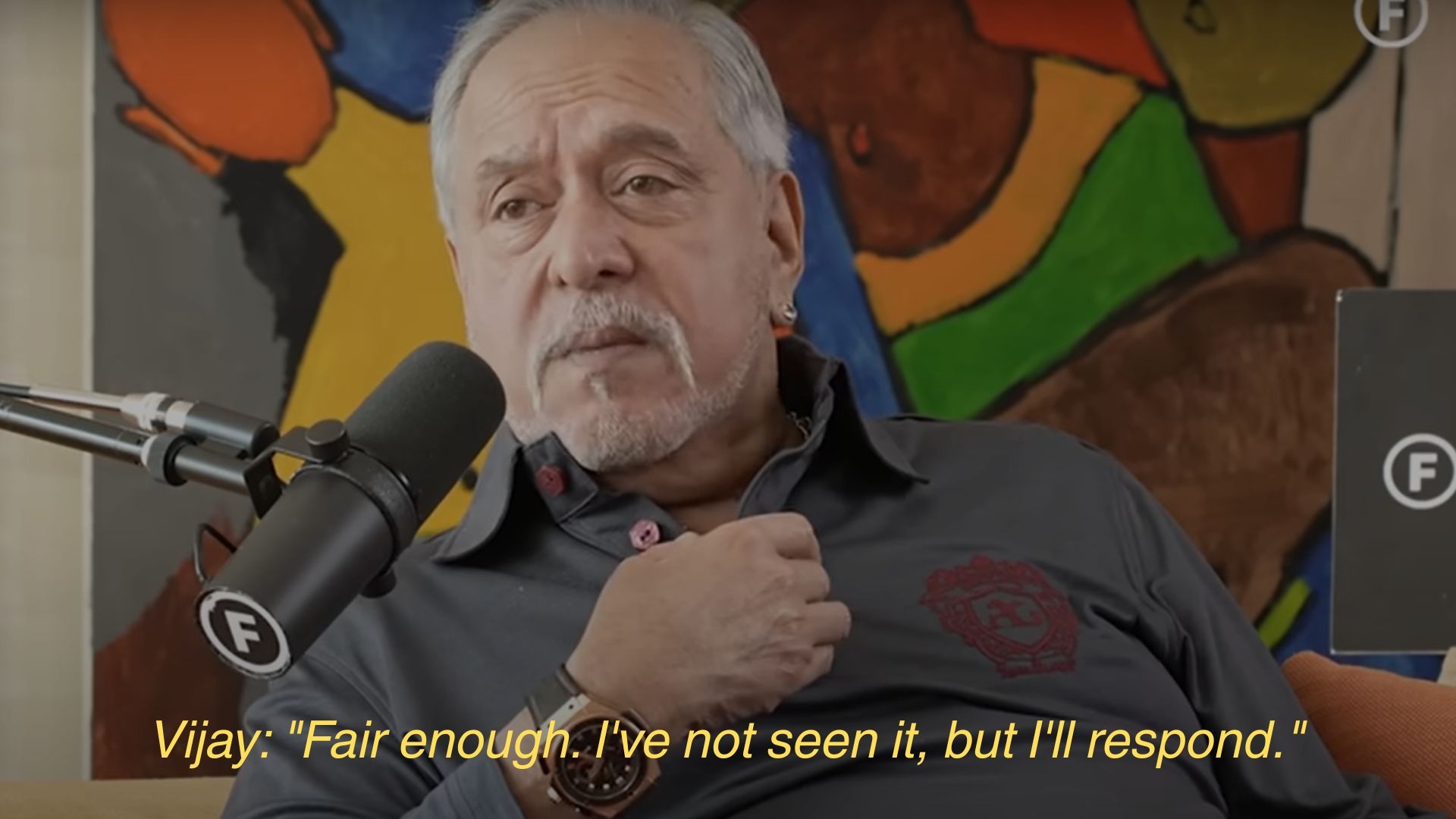 An older man speaks into a microphone during an interview, wearing a collared shirt. Subtitles read: "Fair enough. I've not seen it, but I'll respond."