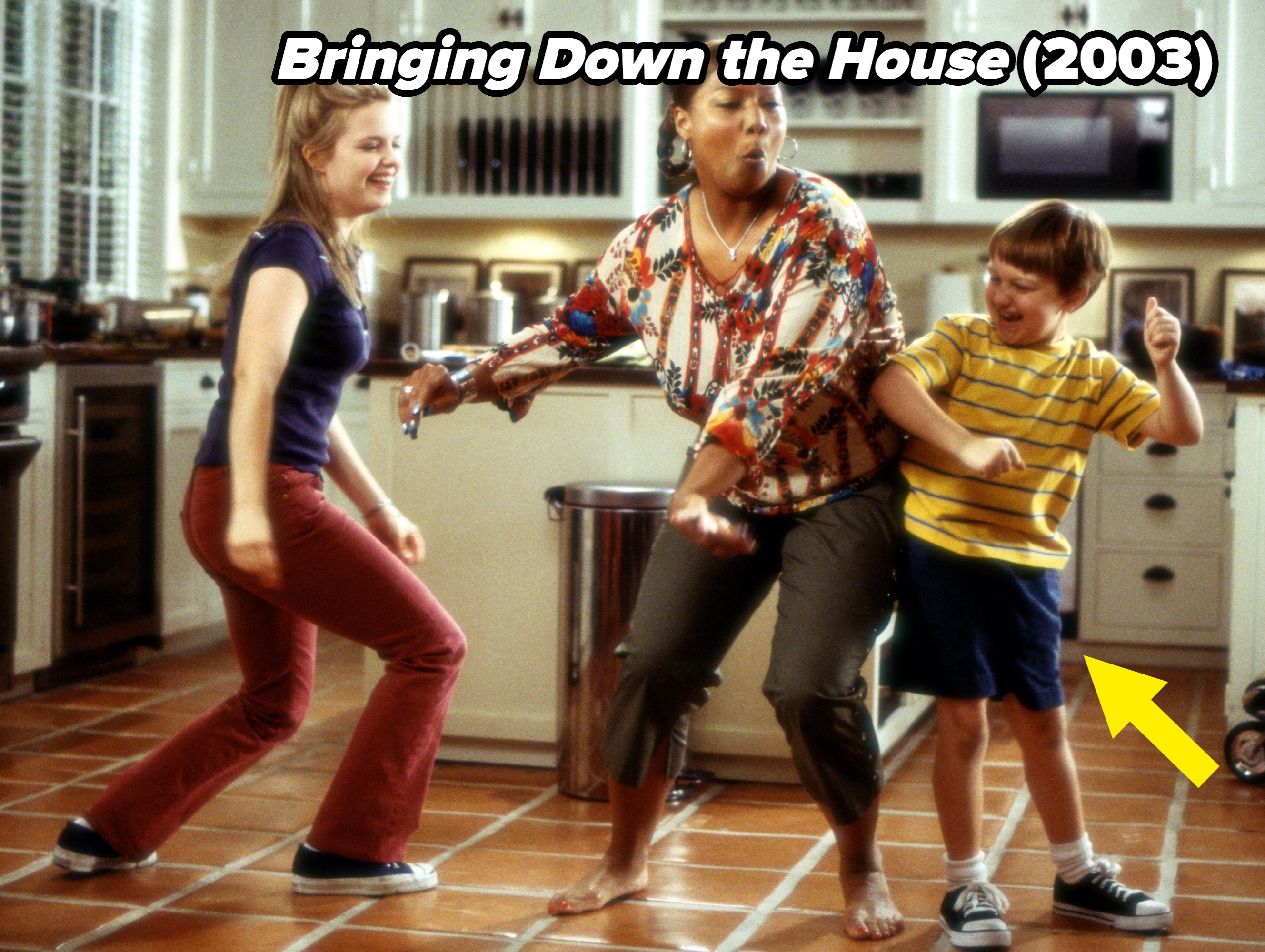 Three people dancing playfully in a kitchen. The person in the center wears a patterned blouse, with others in casual clothes, smiling and having fun