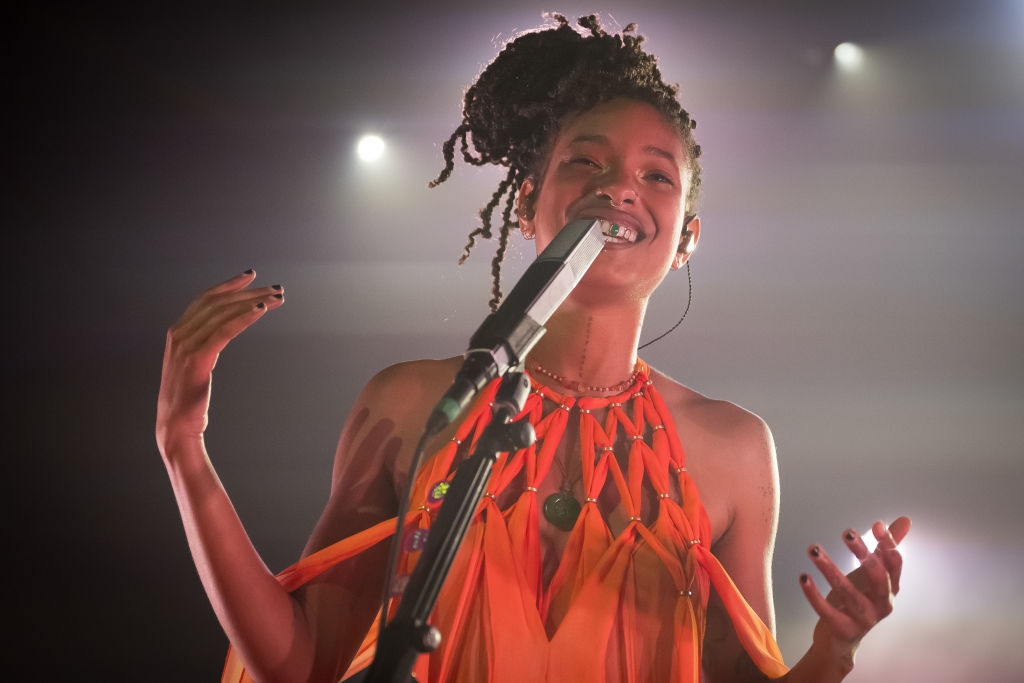 Singer performing passionately on stage, wearing a flowing, artistic halter dress, holding a microphone, with bright stage lights behind