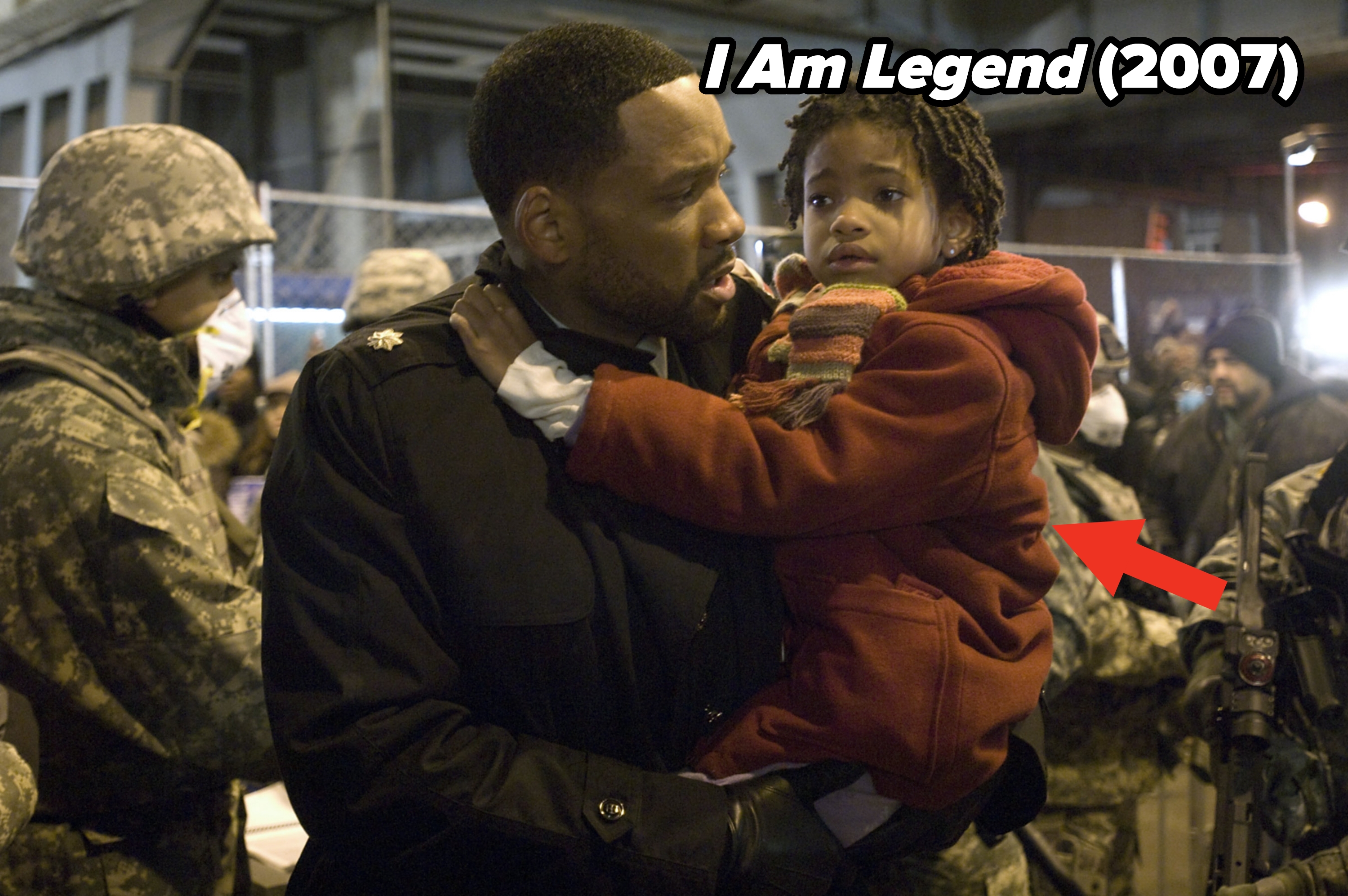 A man in a military-style coat holds a young child in a crowd. Soldiers and fencing are visible in the background