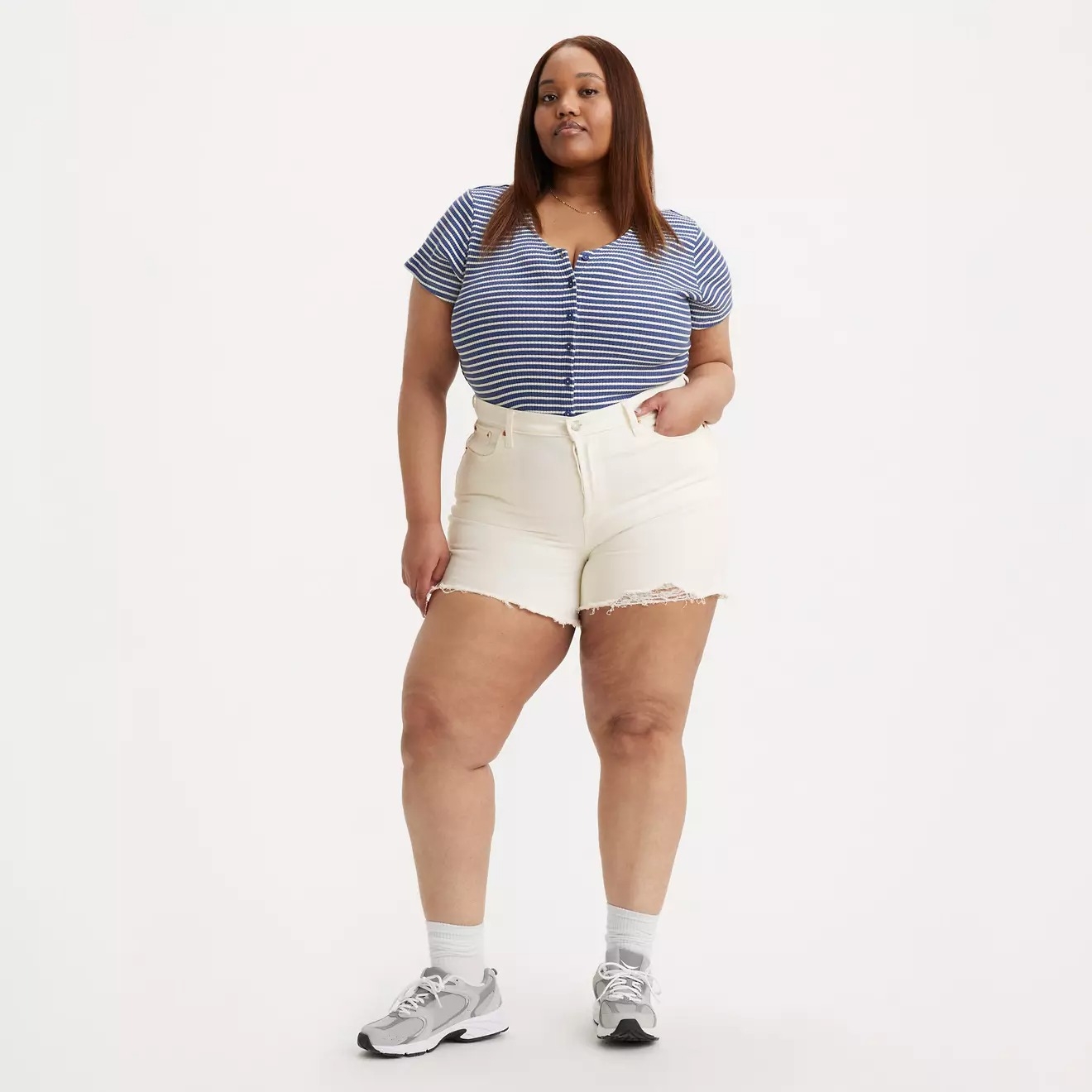 Person wearing a striped top, cream shorts, and sneakers standing confidently on a plain background