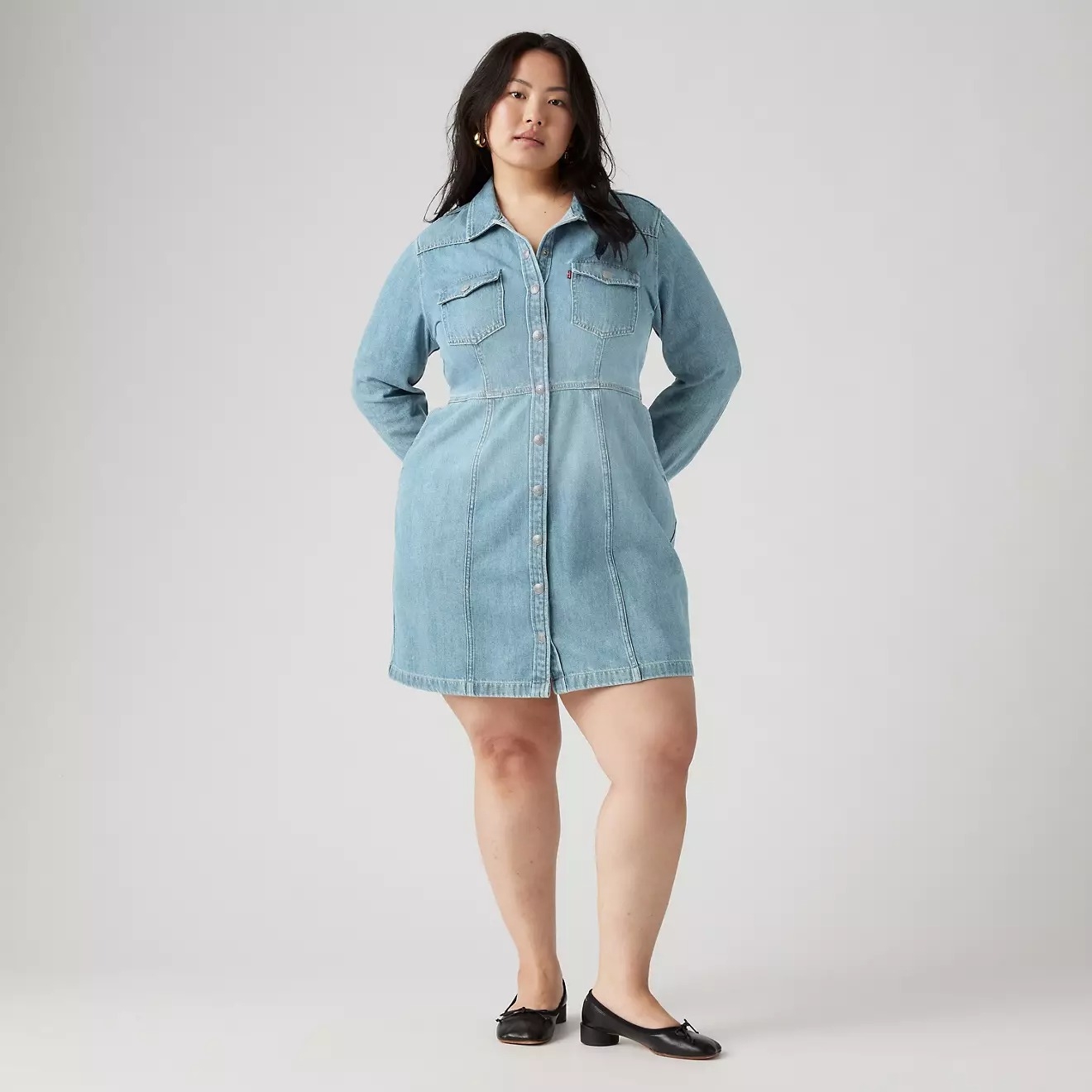 A person models a casual denim dress with a button-front design and long sleeves. They pair it with simple black flats