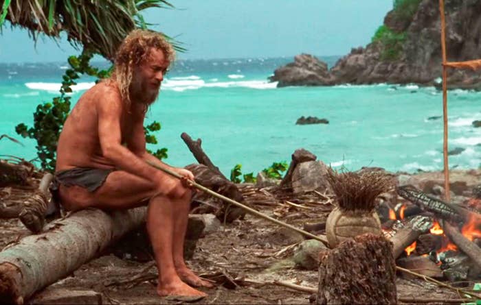 A bearded man in tattered clothing sits on a log near a campfire on a deserted beach, gazing thoughtfully at the flames