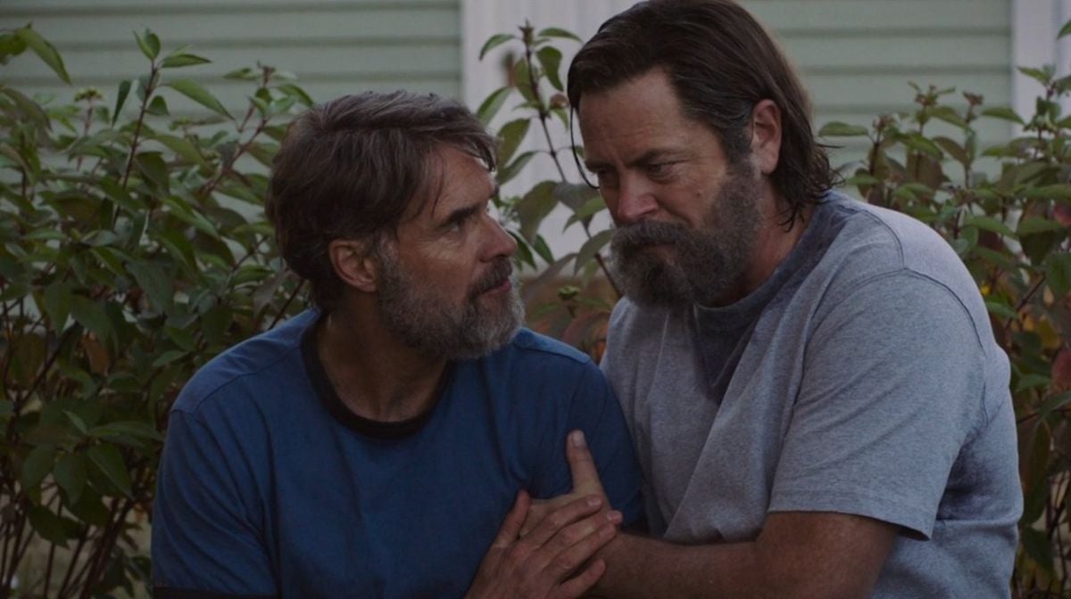 Two men with serious expressions hold hands and appear engaged in an emotional moment, sitting outdoors surrounded by greenery