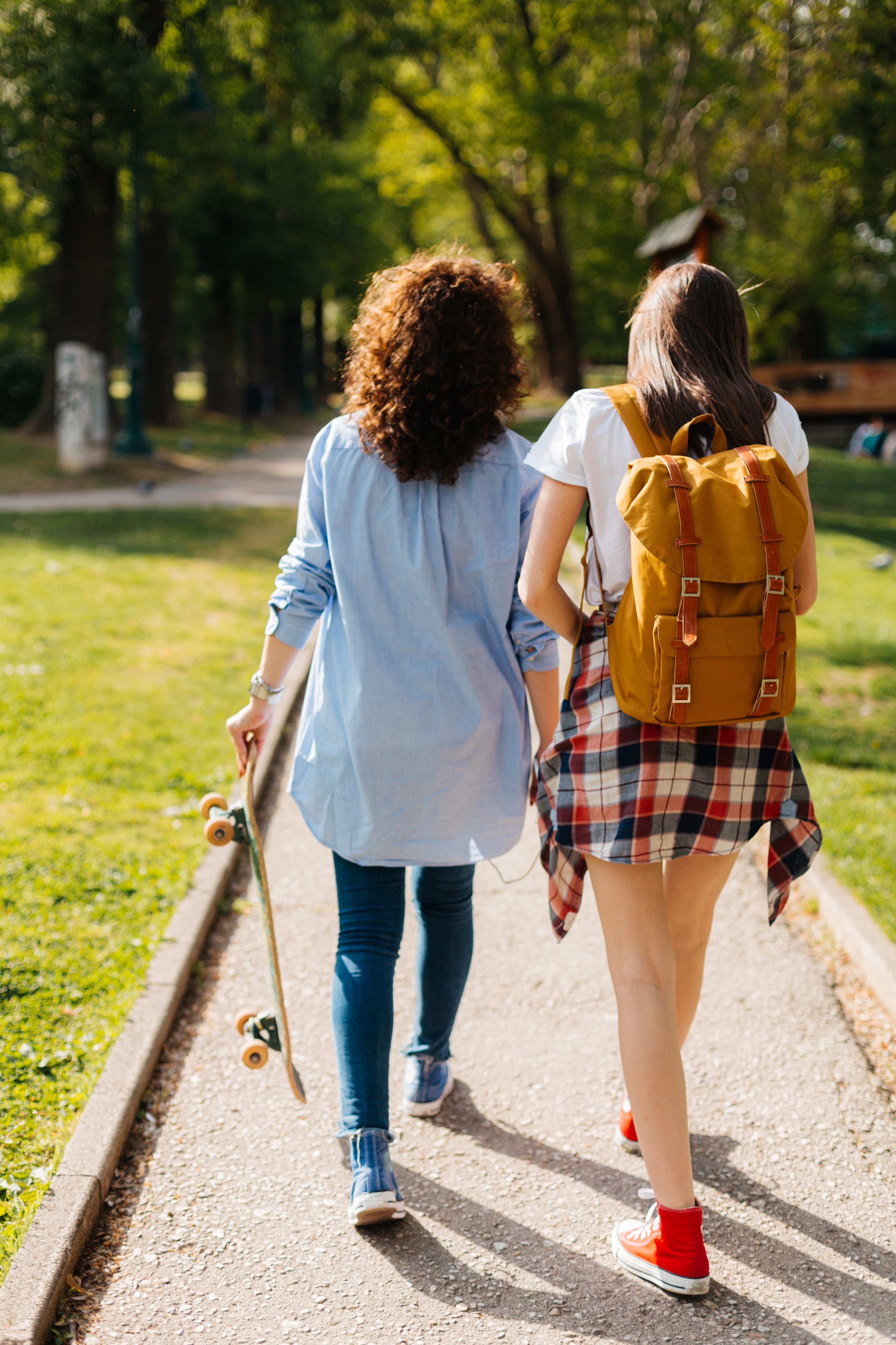 To personer som går på en sti i en park. En holder et skateboard. Begge bruker uformelle klær og ryggsekker. Trær og gress stiller stien